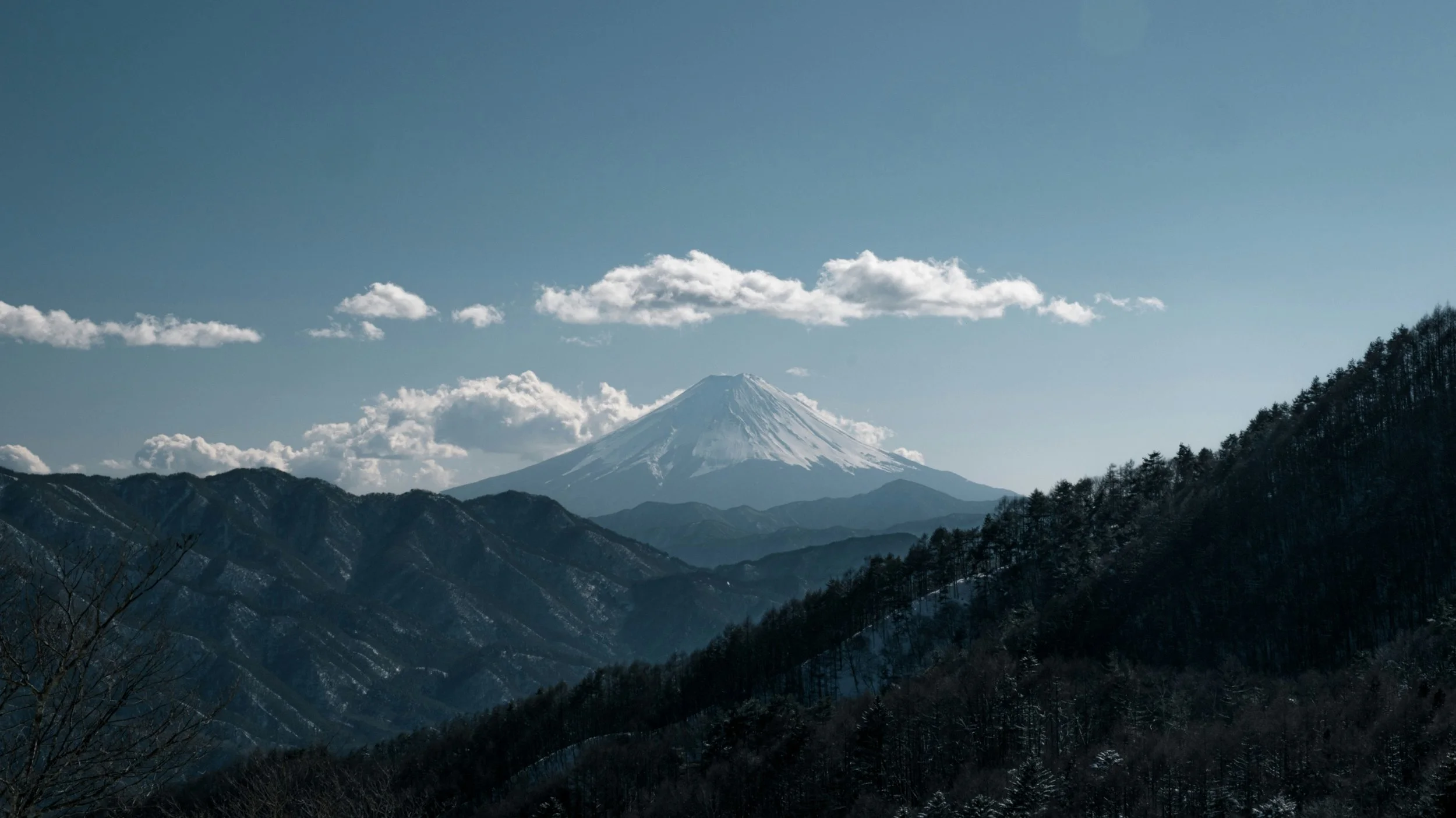 Snow-capped Mount Fuji against a blue sky with clouds over rugged mountain terrain in winter.