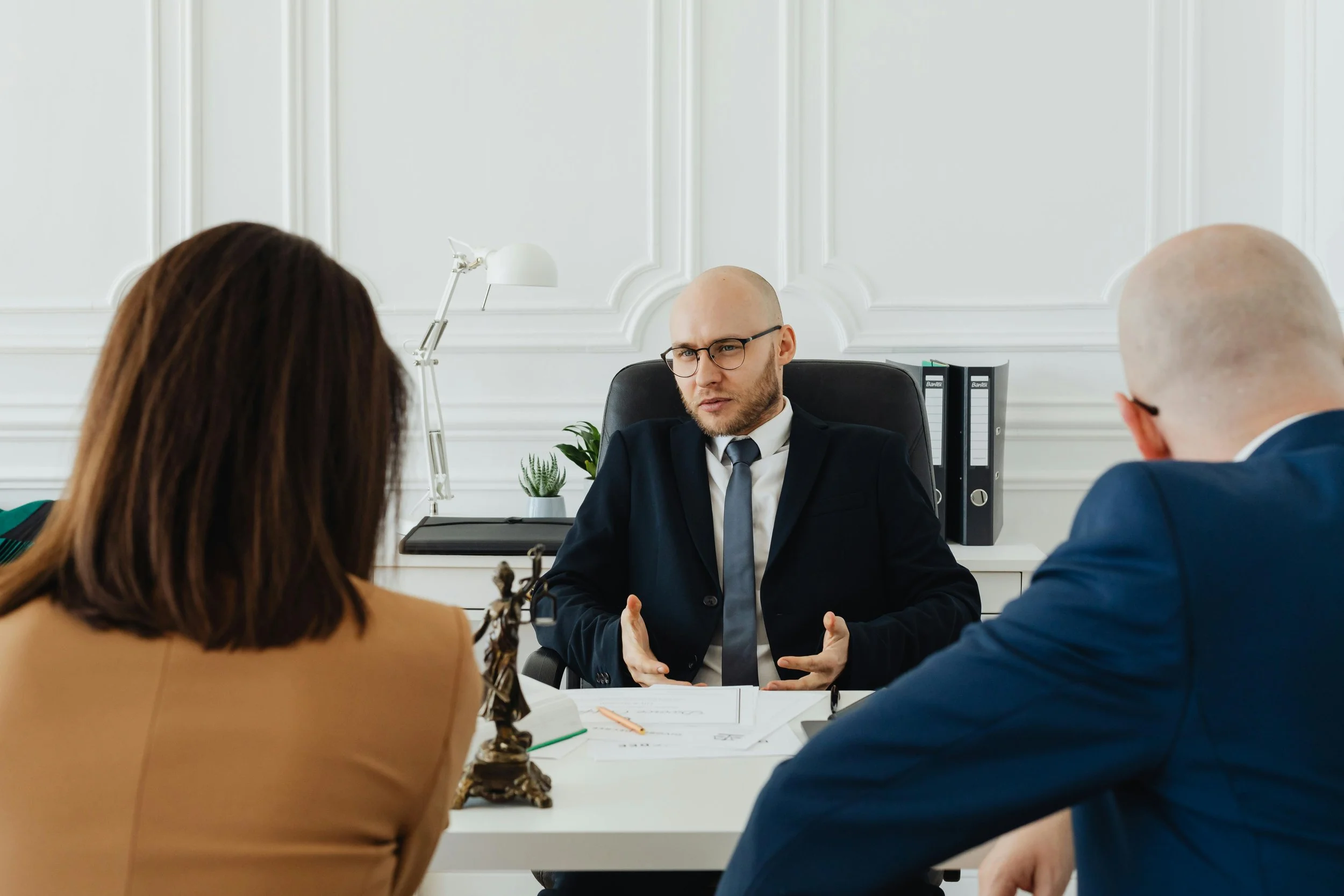 A professional office scene showing a man in a suit speaking to two people across a white desk.