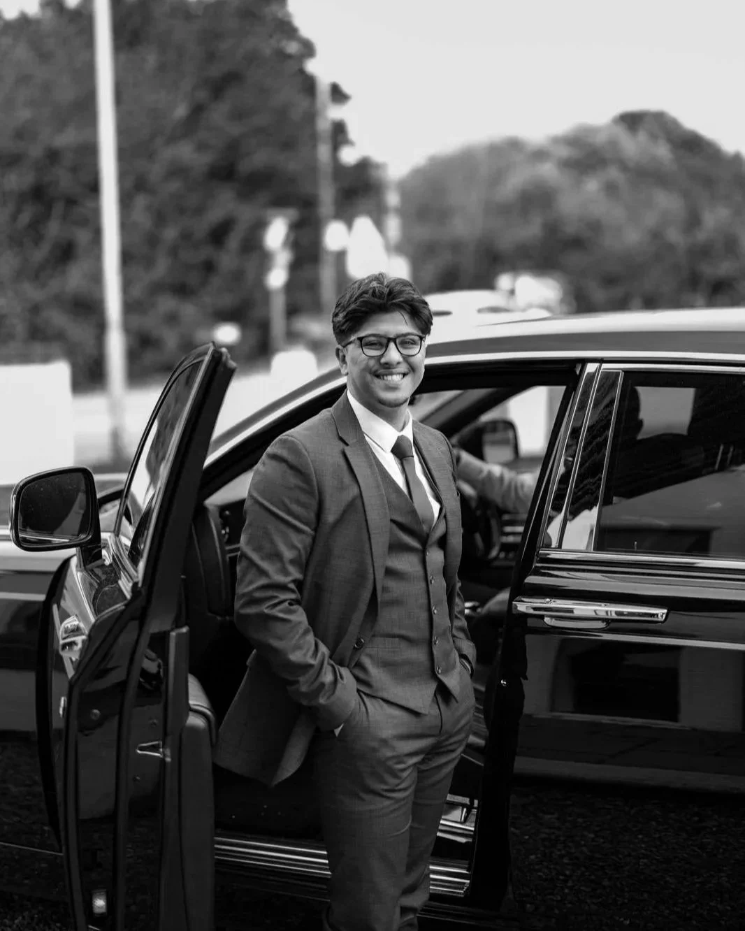 A smiling man in a suit standing outside a car with the door open, wearing glasses, with a background of trees and power lines.