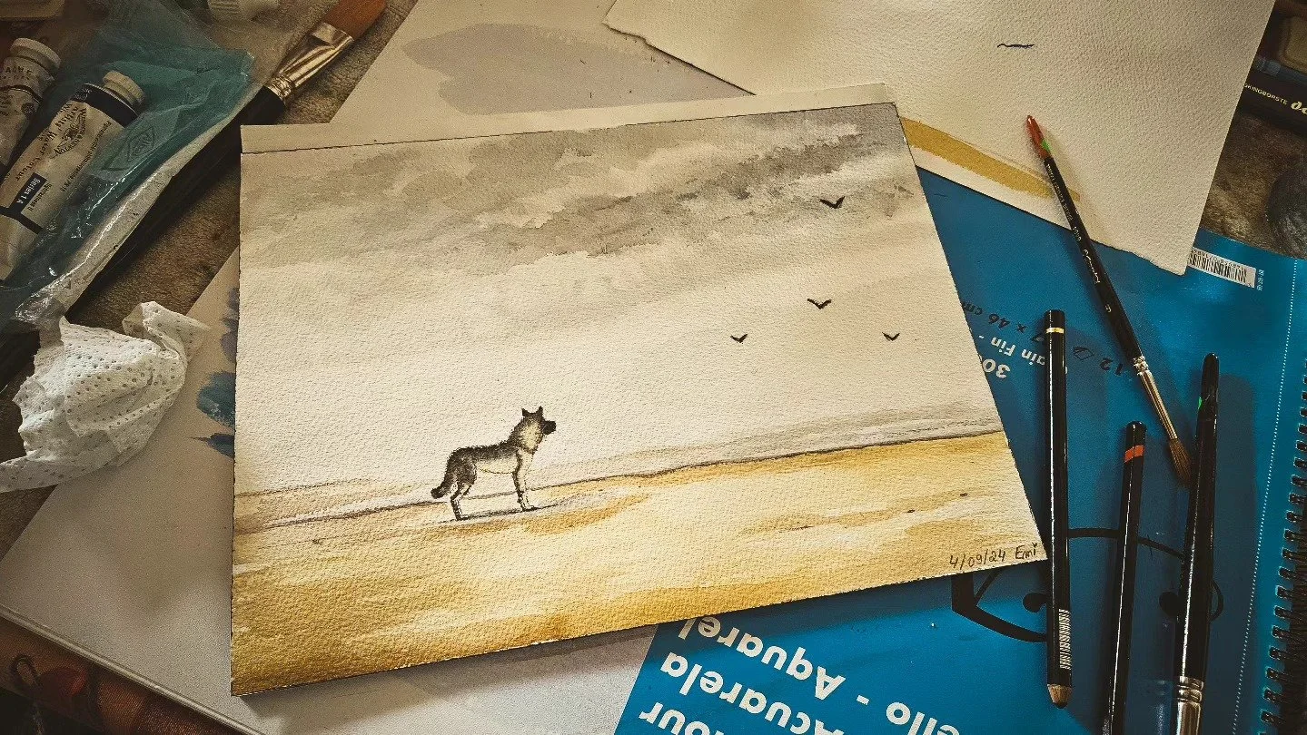 Une aquarelle représentant un loup dans un paysage désertique avec un ciel gris nuageux, quelques oiseaux volants et un fond beige et gris, avec des pinceaux et des tubes de peinture à l'écran.