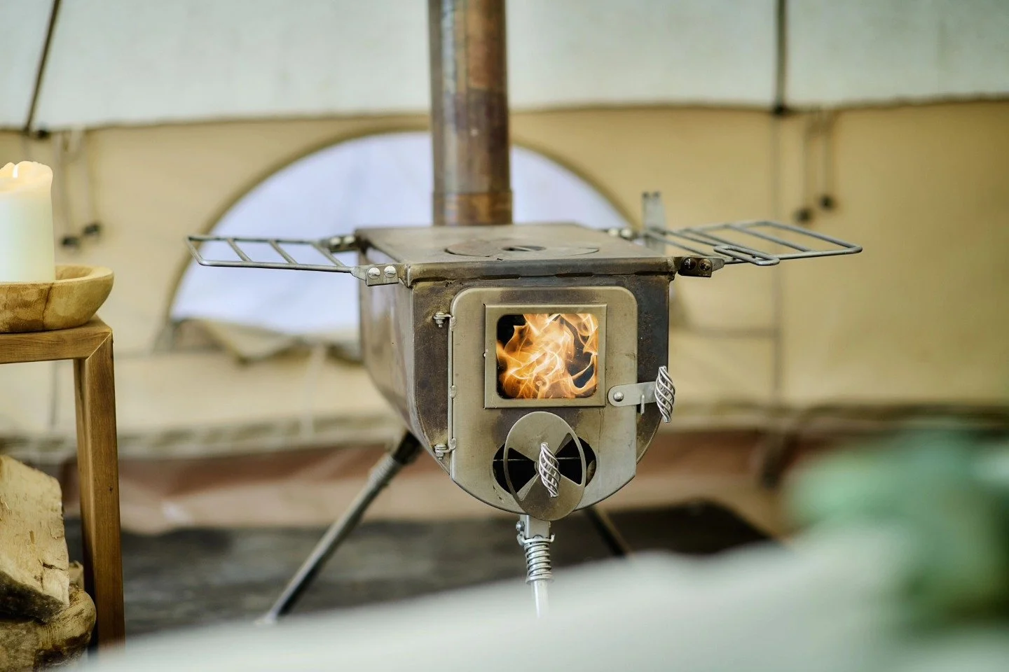 Un poêle à bois métallique avec feu visible à l'intérieur, placé dans une tente ou un abri, avec une structure en métal et un conduit de fumée en position verticale.