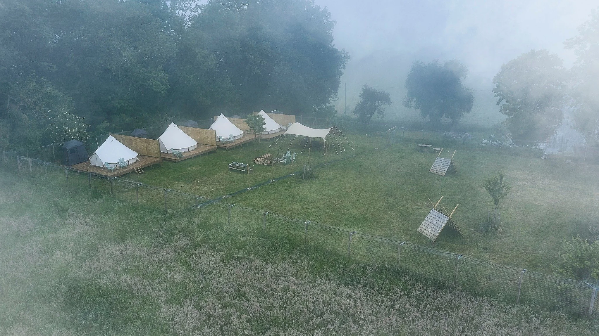 Tentes blanches sur une plateforme en bois, entourées de forêt et de brume matinale.