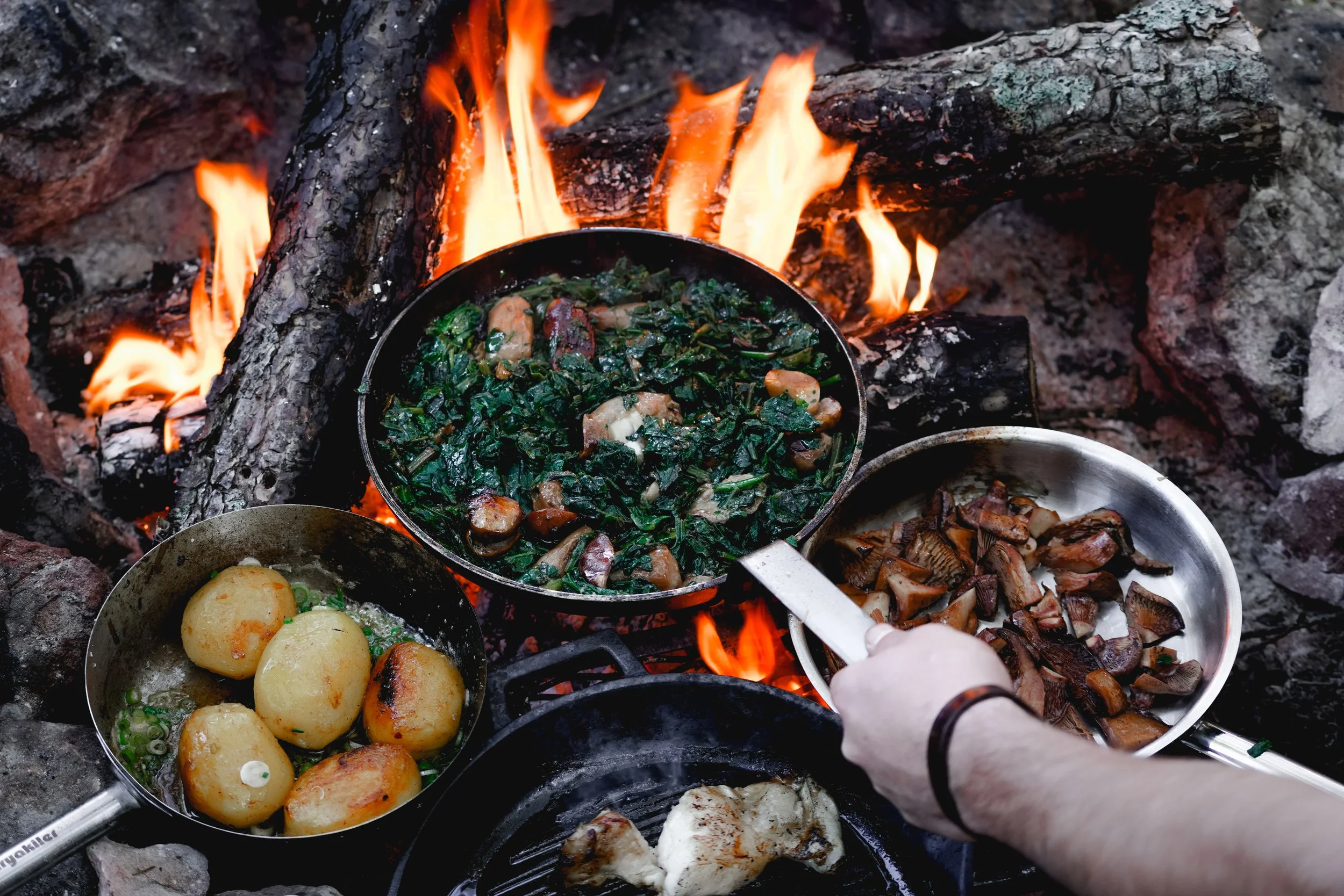Cuisson de différents aliments dans des poêles sur un feu de camp en plein air, avec des grattons, pommes de terre, champignons et légumes verts.