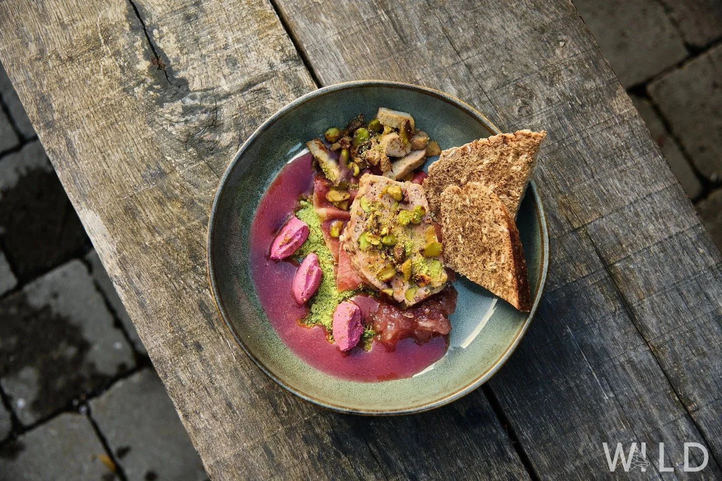 Assiette avec un plat de dégustation comprenant des morceaux tapissés de pistaches, du pain grillé, et une sauce violette sur une table en bois rustique.