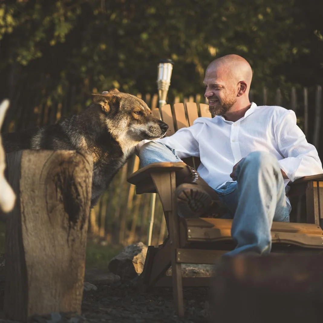 Un homme souriant assis dans une chaise en bois, touchant doucement un chien qui se repose sur un tronc d'arbre, dans un cadre extérieur. La scène est chaleureuse, avec la lumière du coucher de soleil et un fond de nature avec des arbres.