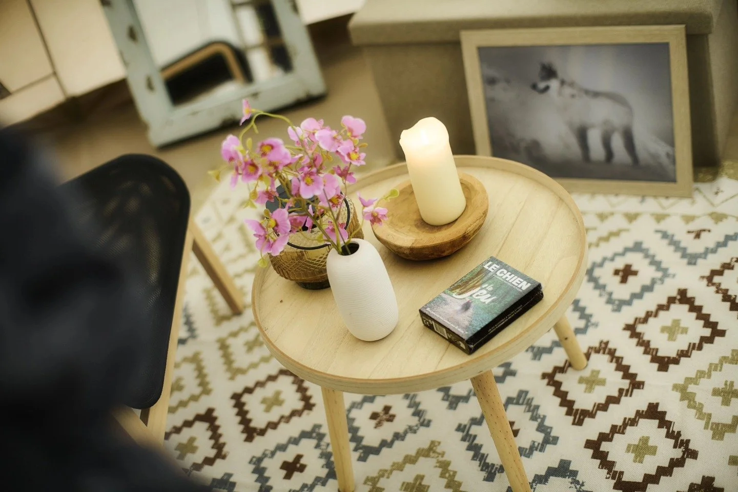 Table en bois claire avec une plante de fleurs roses dans une vase blanche, une bougie blanche dans un porte-bierre en bois, un livre intitulé 'Le Chien' et une photo encadrée noir et blanc d'un chien sur un tapis de motifs géométriques en noir, blanc et beige.
