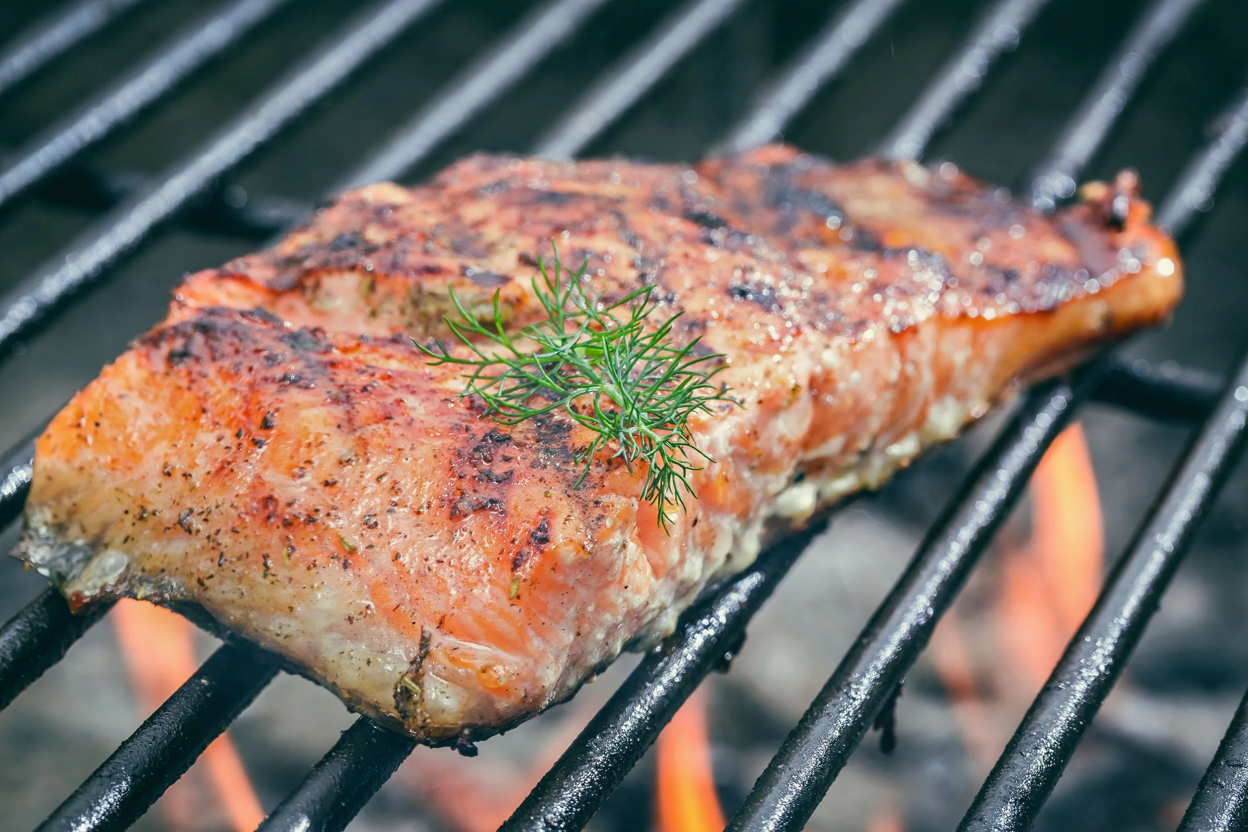 Un filet de saumon grillé avec une branche de dille, sur une grille de barbecue avec des flammes en dessous.