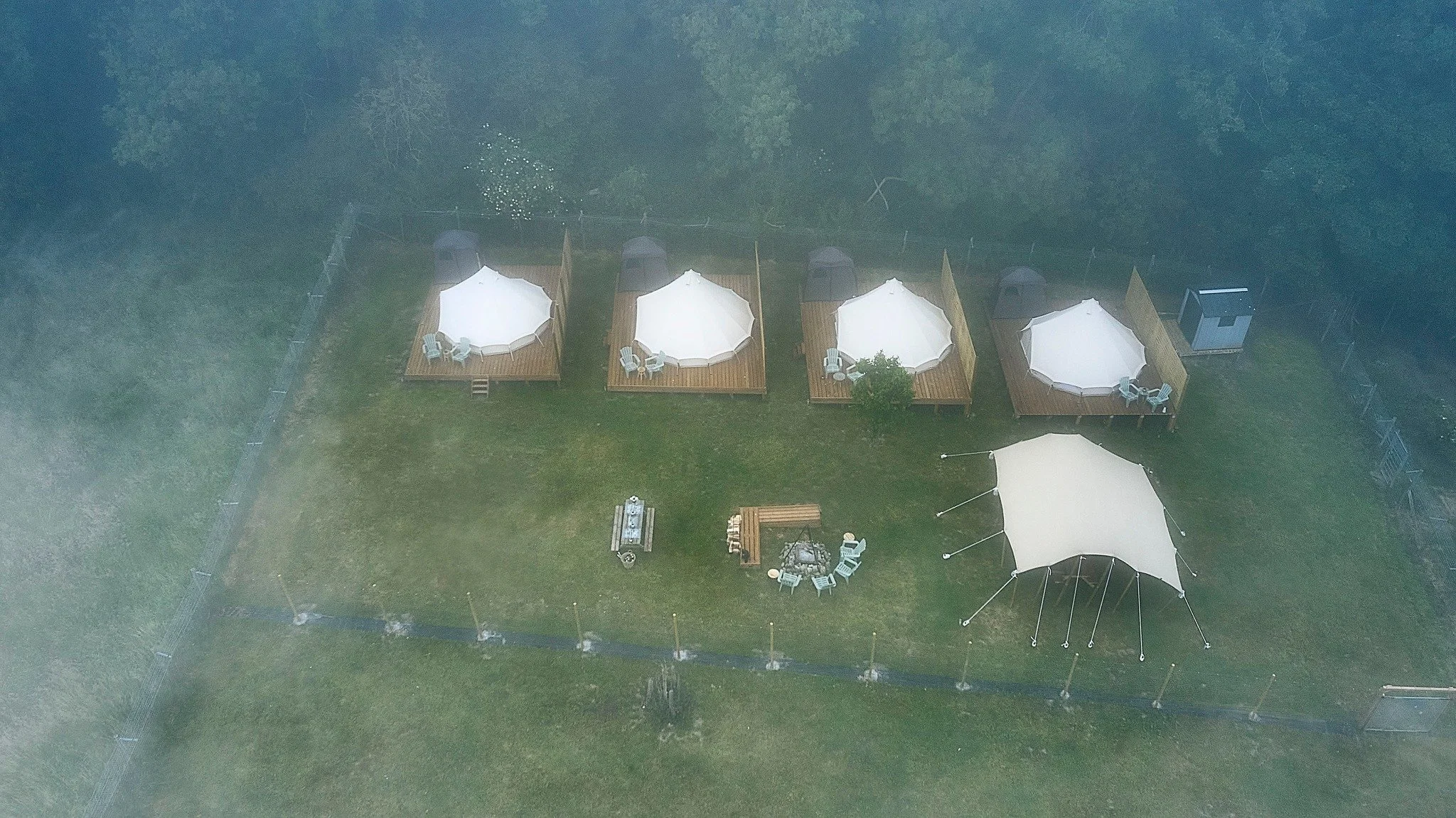 Vue aérienne d'un espace en plein air avec quatre cabanes en bois, chacune avec une grande parasol blanc et mobilier de terrasse, entouré d'une clôture en bois et de végétation dense en arrière-plan.
