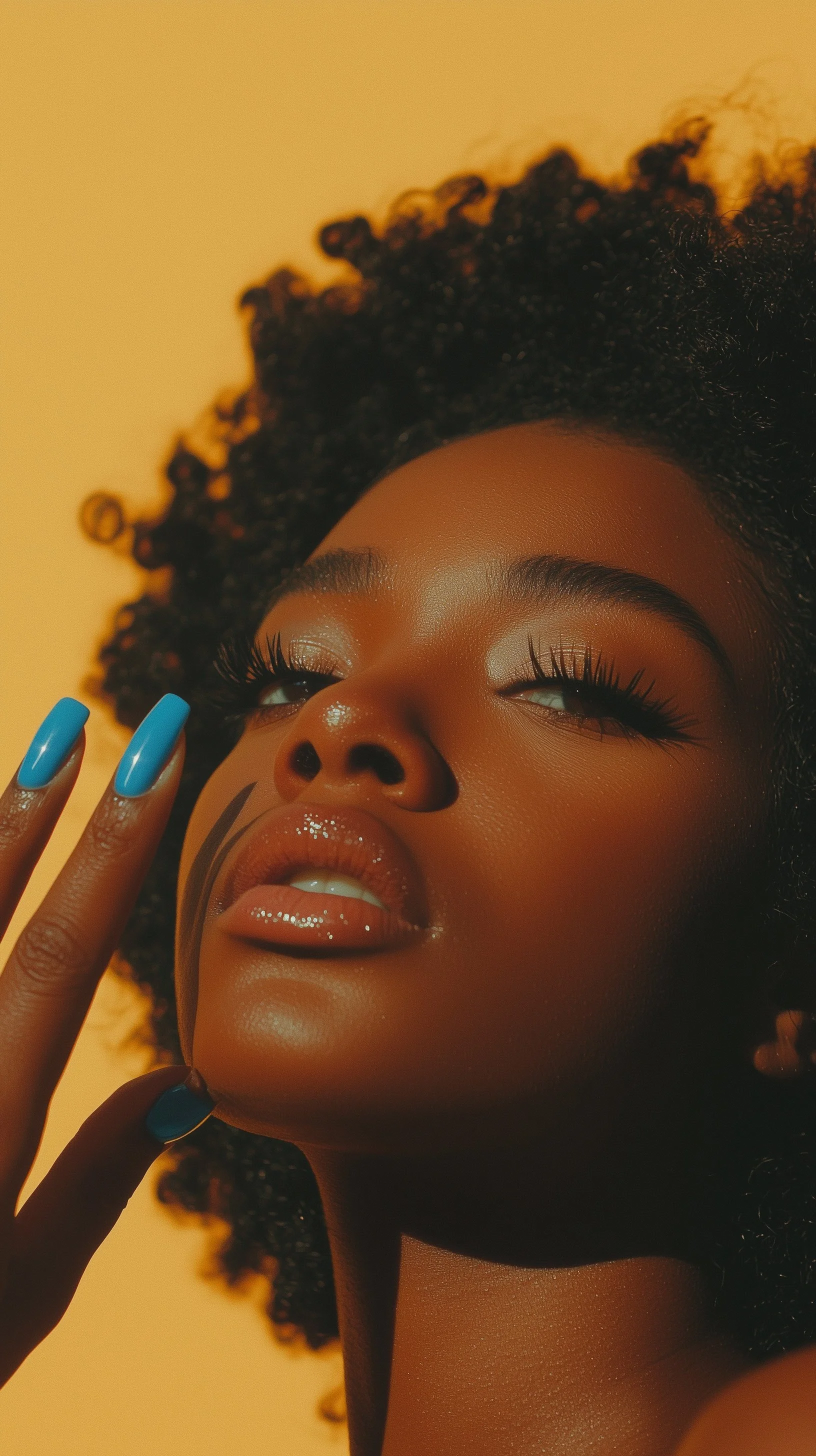 Close-up portrait of a Black woman with curly hair, wearing glossy nude lipstick, shimmery eye makeup, and blue nail polish, posed with her eyes partially closed and touching her face.