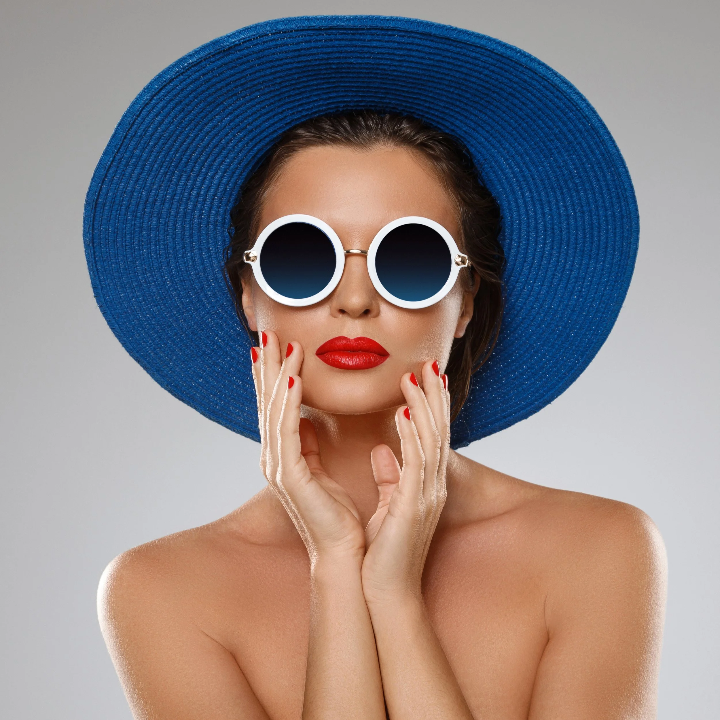 A woman wearing a large blue sunhat, white round sunglasses with dark lenses, and red lipstick. She has red nail polish and is holding her face with her hands. The background is plain gray.
