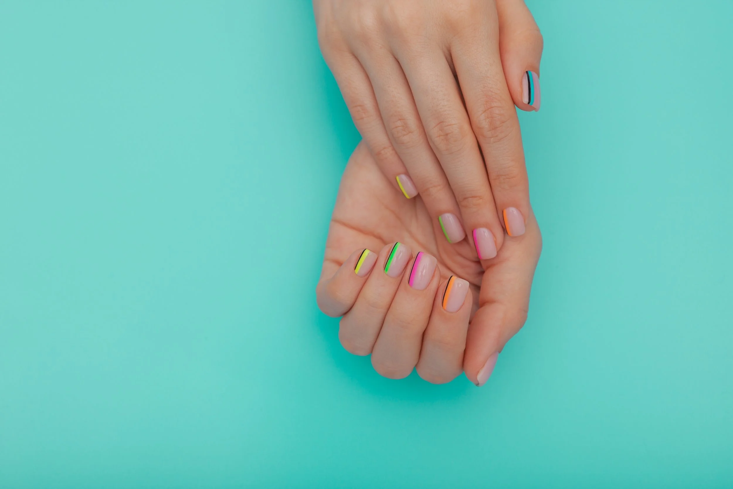 Close-up of hands with colorful French manicure nails in various colors against a turquoise background.