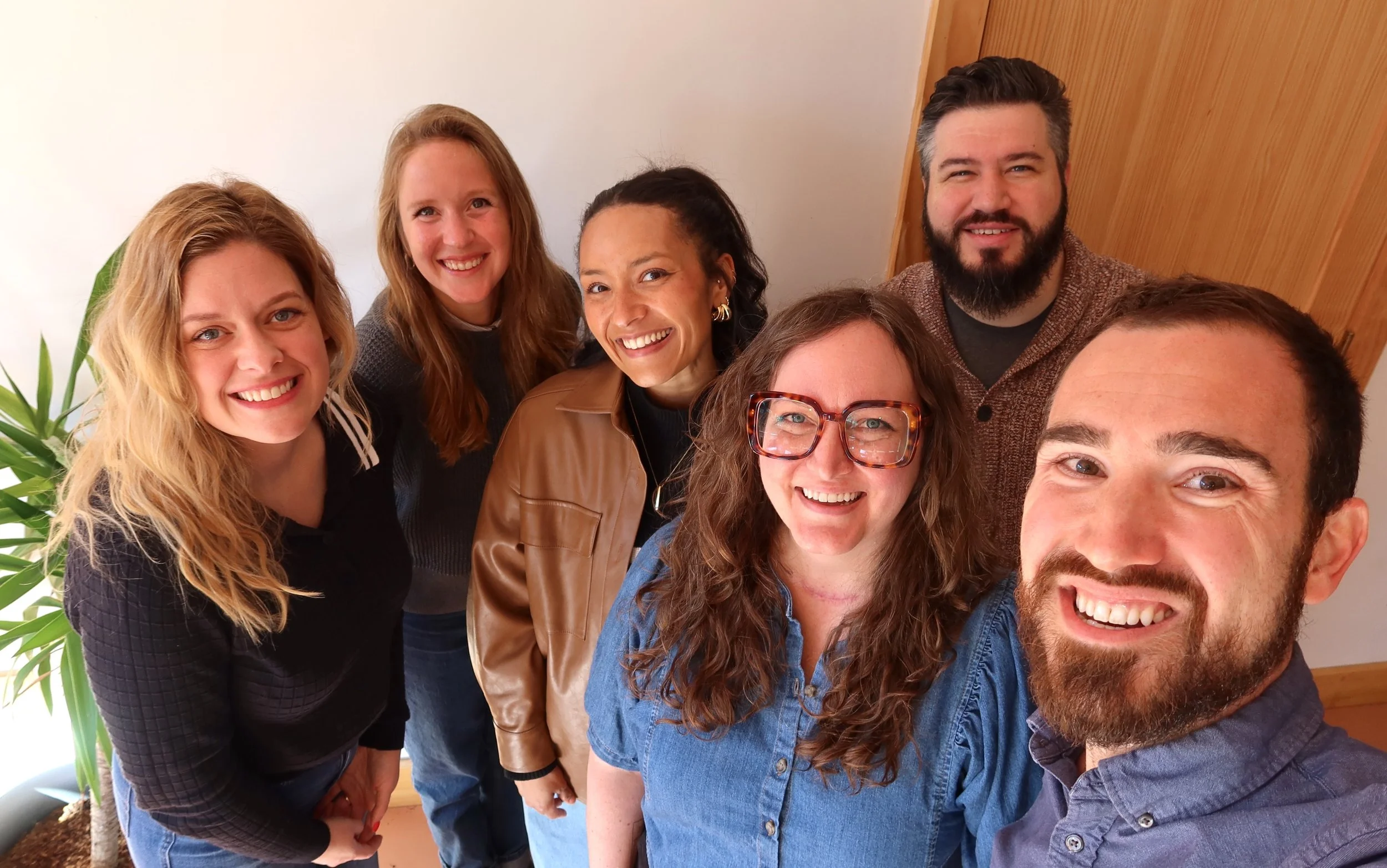 Group of seven people taking a selfie indoors, smiling and looking at the camera, with a large green plant and wooden wall in the background.