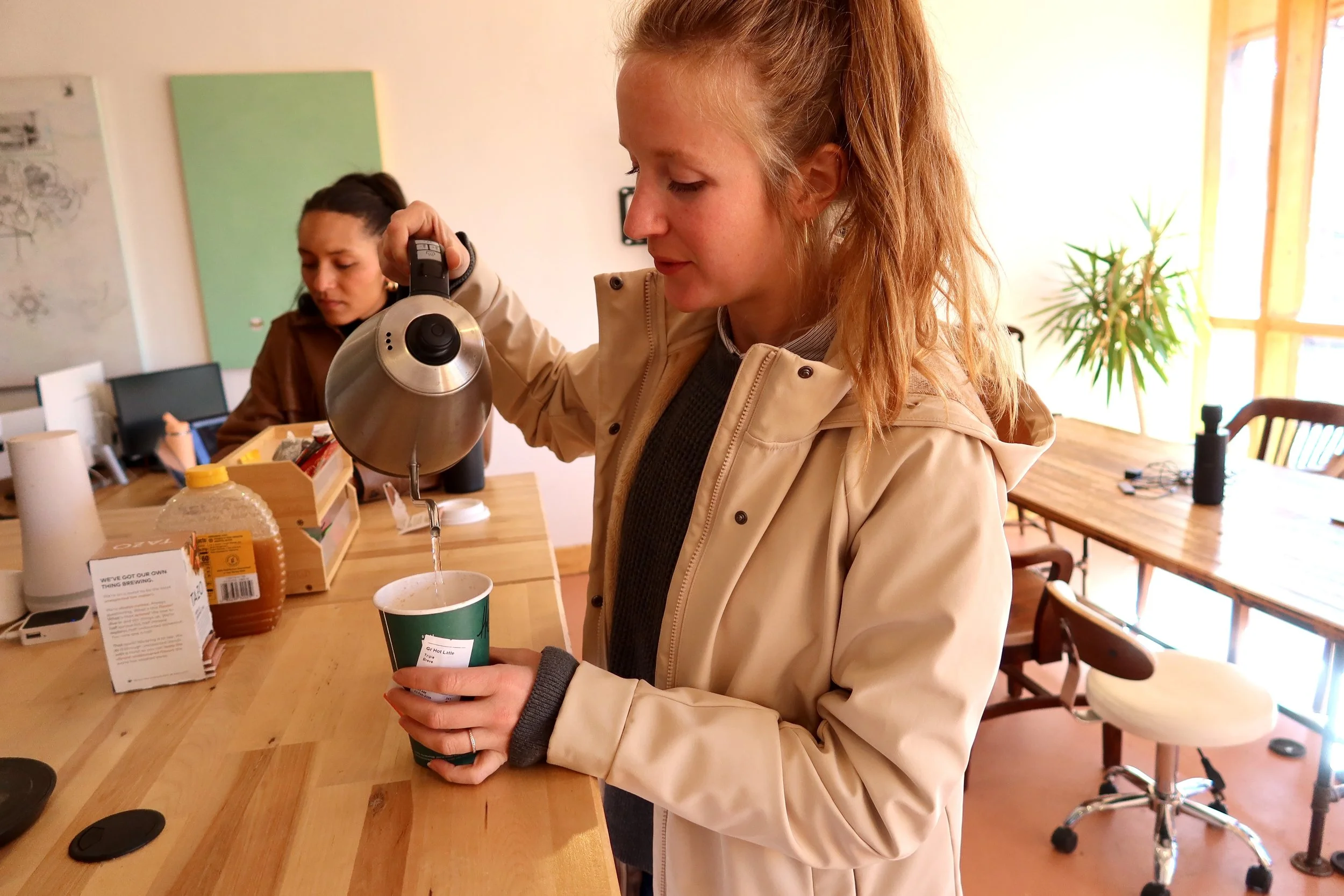 A woman pouring hot water from a kettle into a green disposable cup at a wooden counter in a cozy, well-lit room with another woman working in the background.