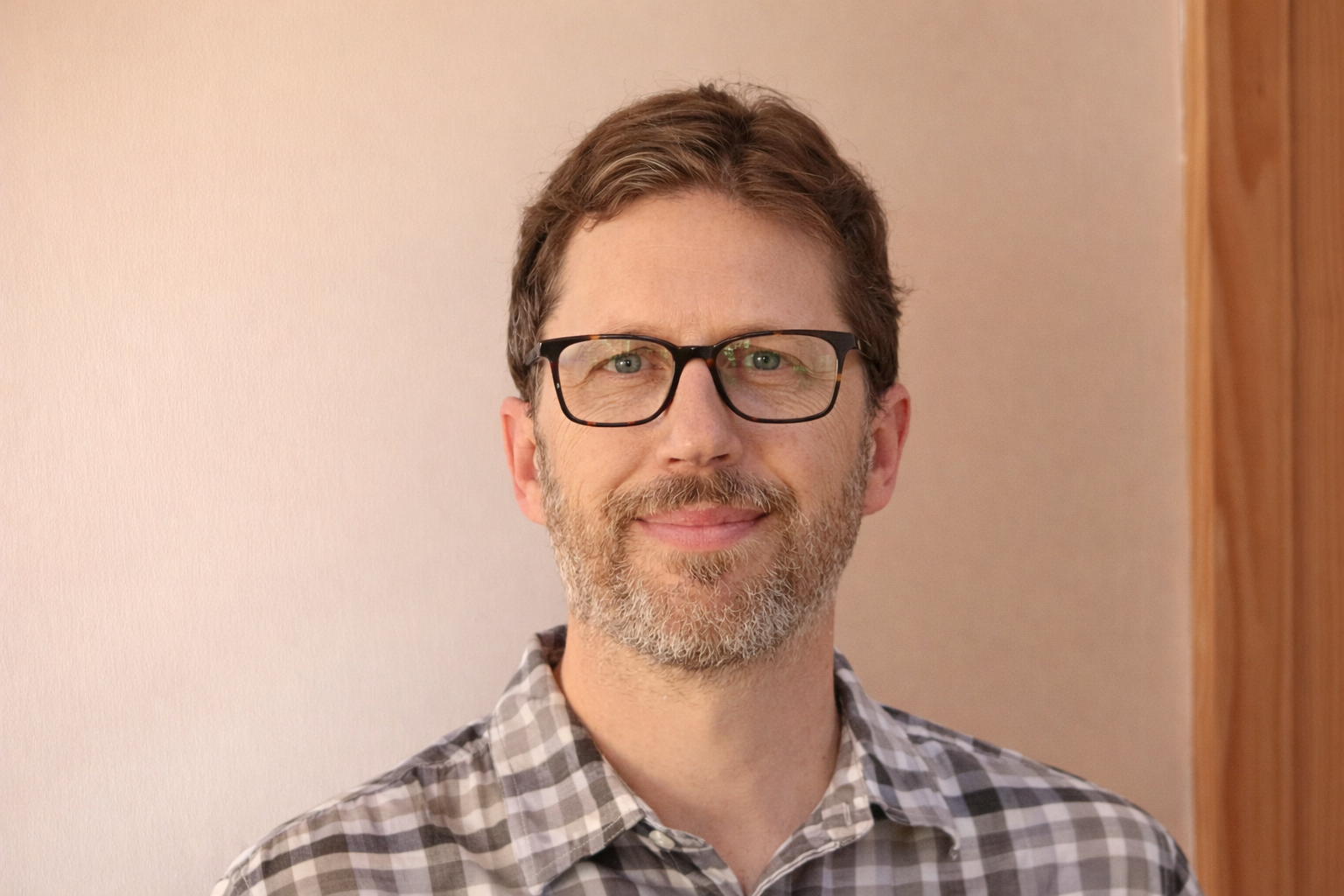 A smiling man with glasses and a beard, wearing a checkered shirt, standing indoors near a wooden wall and a plain wall.