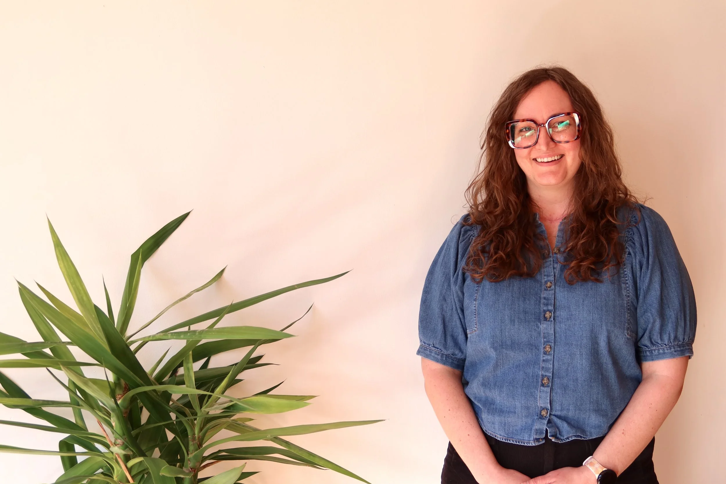 A woman with long curly brown hair wearing glasses and a denim shirt standing next to a green plant in front of a plain beige wall.