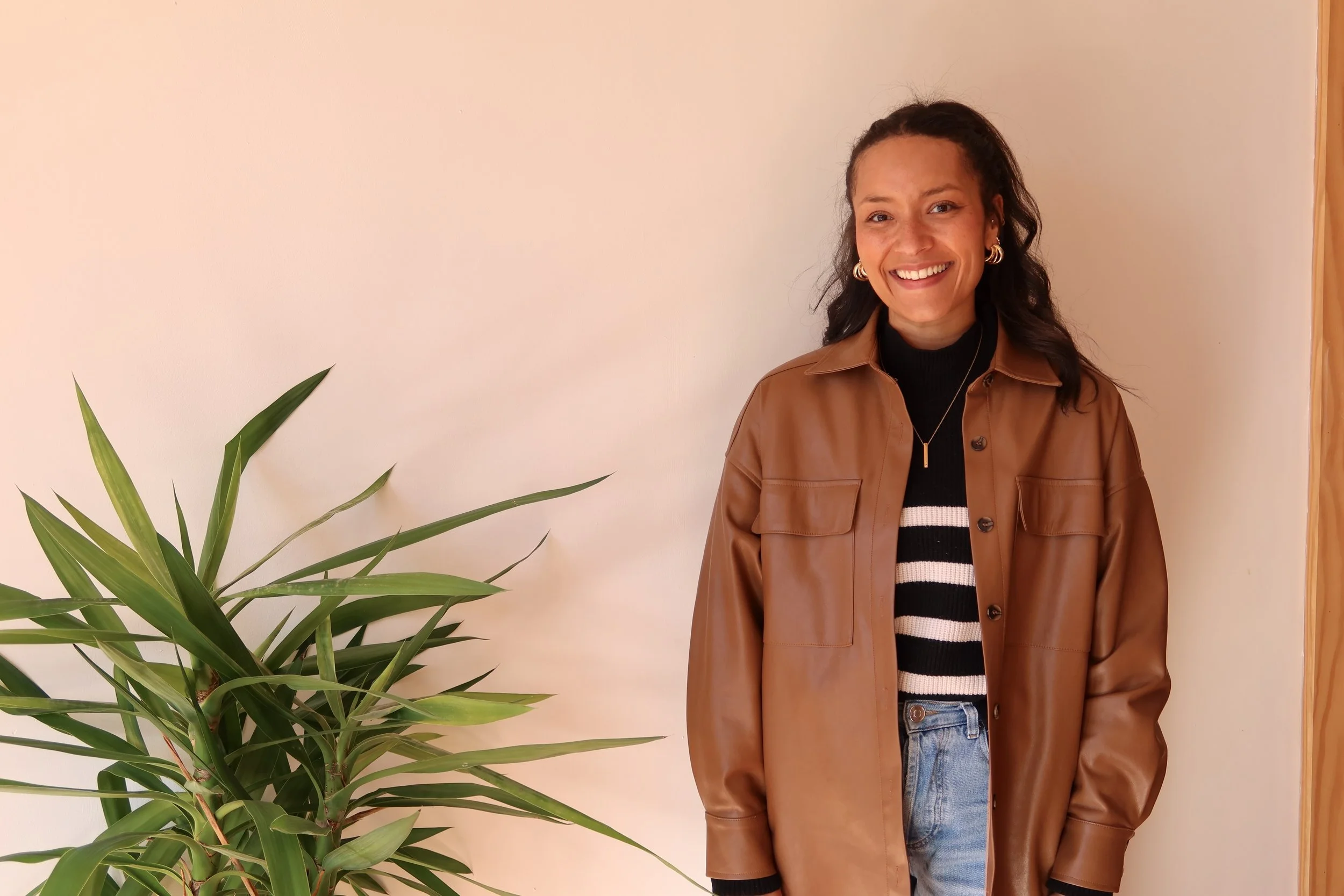 Smiling woman with dark hair wearing a brown leather jacket, black and white striped sweater, and jeans, standing next to a large green plant against a beige wall.