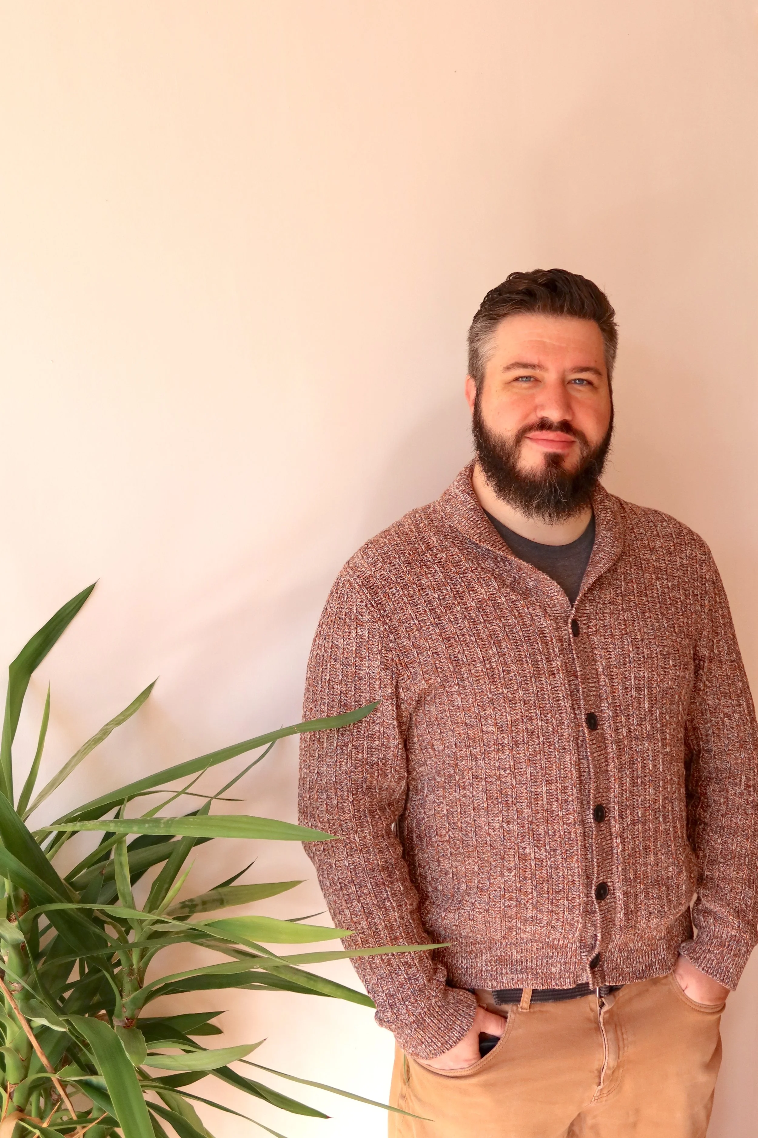 A man with a beard and short hair standing next to a large potted plant inside a room with a plain light-colored wall.