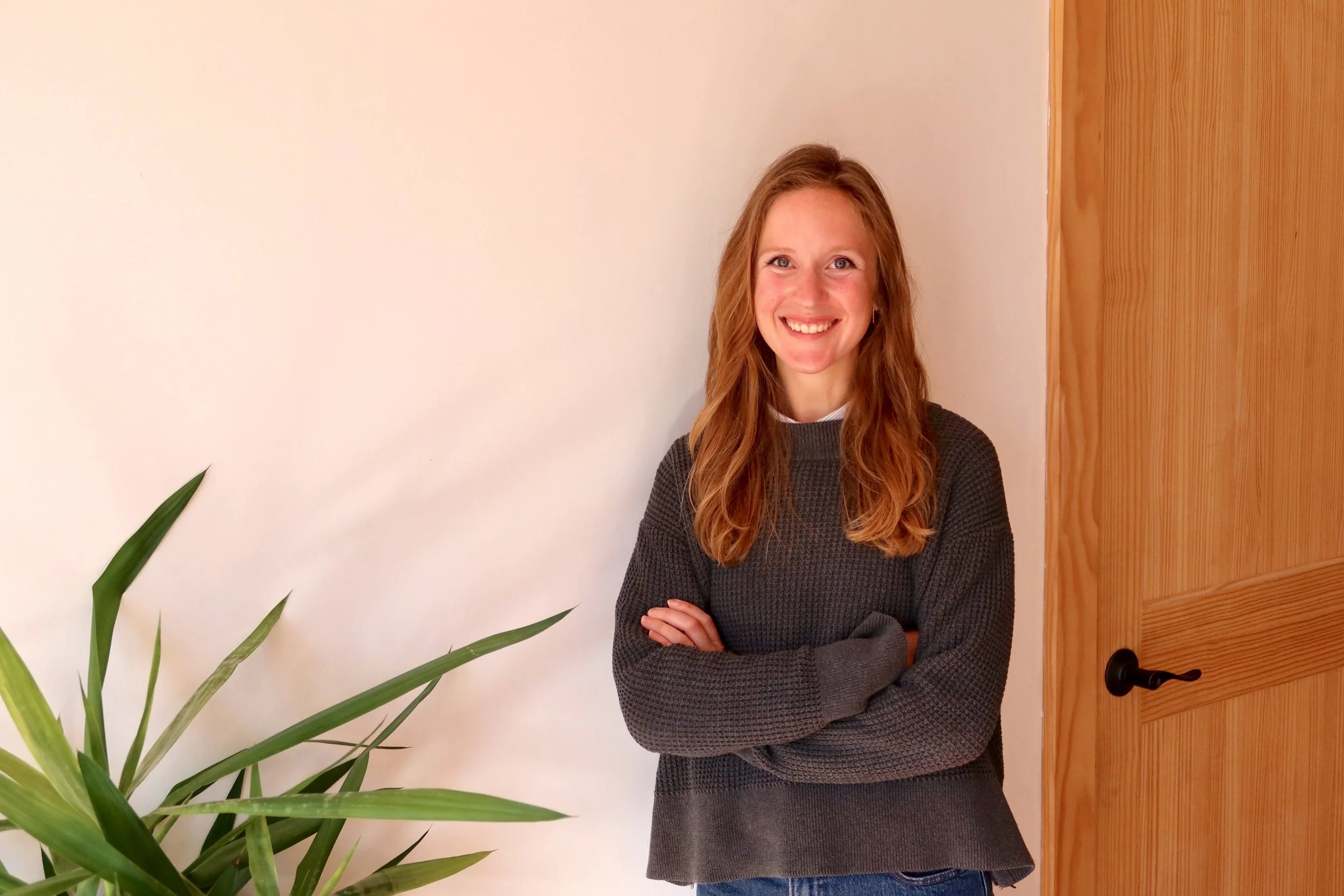 A woman with long blond hair, wearing a black sweater, standing with folded arms and smiling, indoors near a wooden door and a green plant.