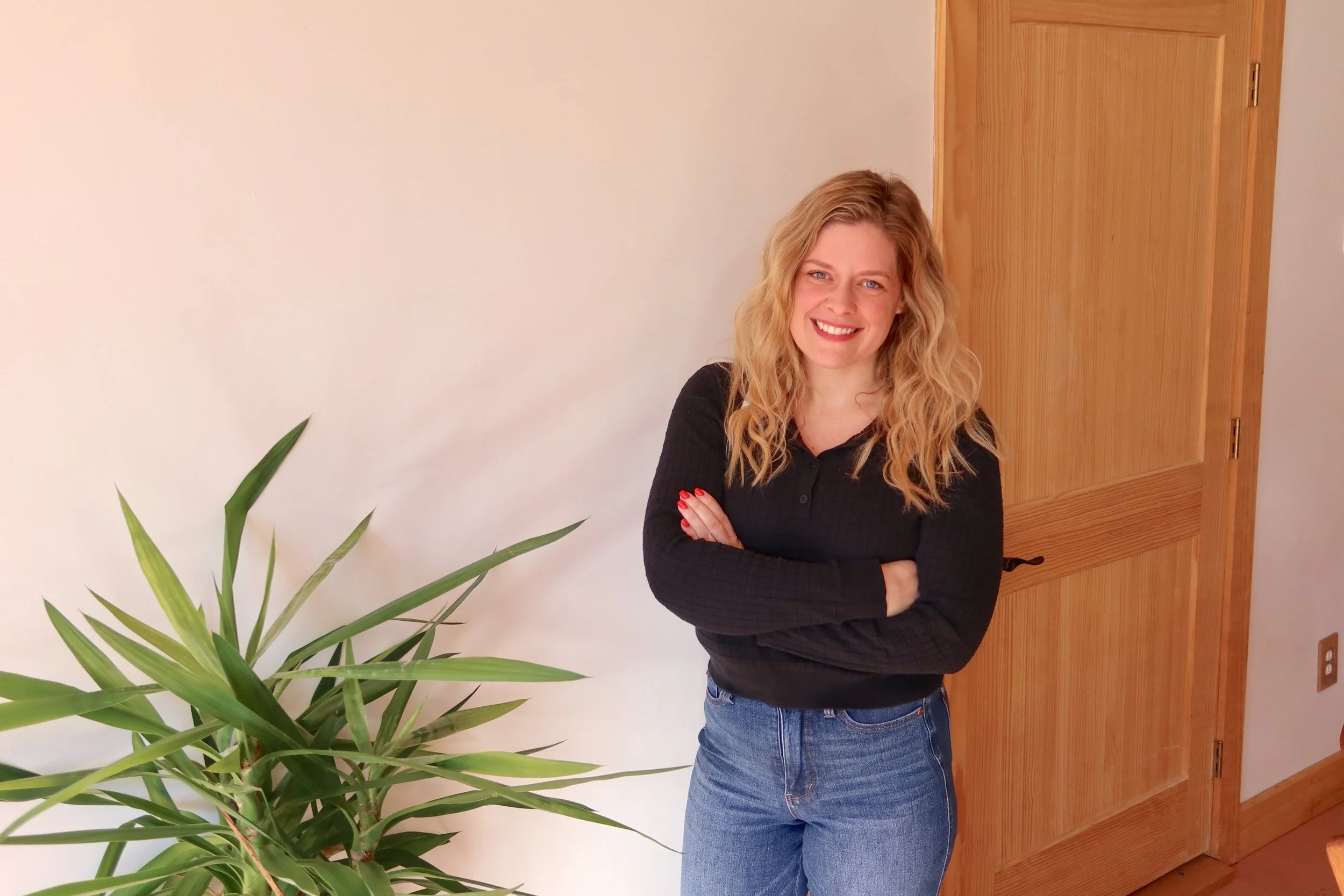 A woman with wavy blonde hair, wearing a black long-sleeve top and blue jeans, standing indoors with her arms crossed, smiling, next to a large green plant, with a wooden door in the background.