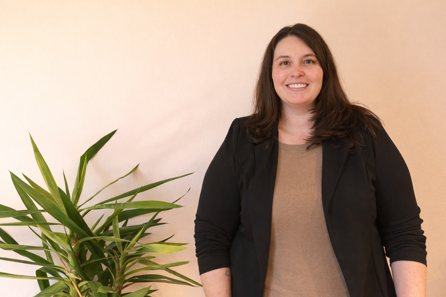 A woman with brown hair smiling, wearing a black blazer over a tan top, standing next to a green plant against a beige wall.