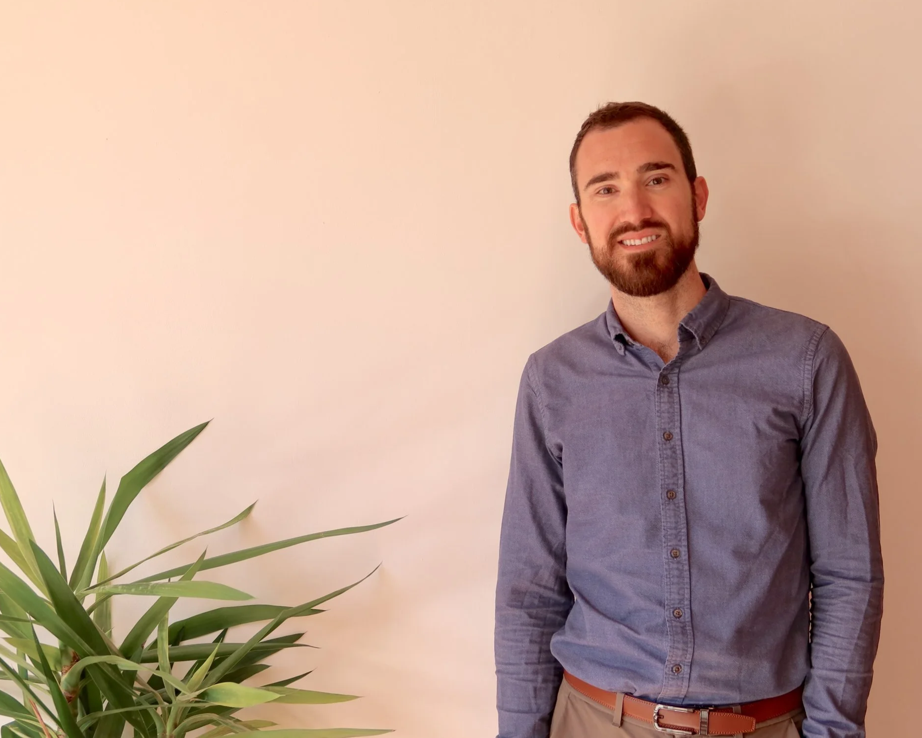 A man with a beard smiling, wearing a blue button-up shirt and khaki pants, standing next to a green plant against a plain white wall.