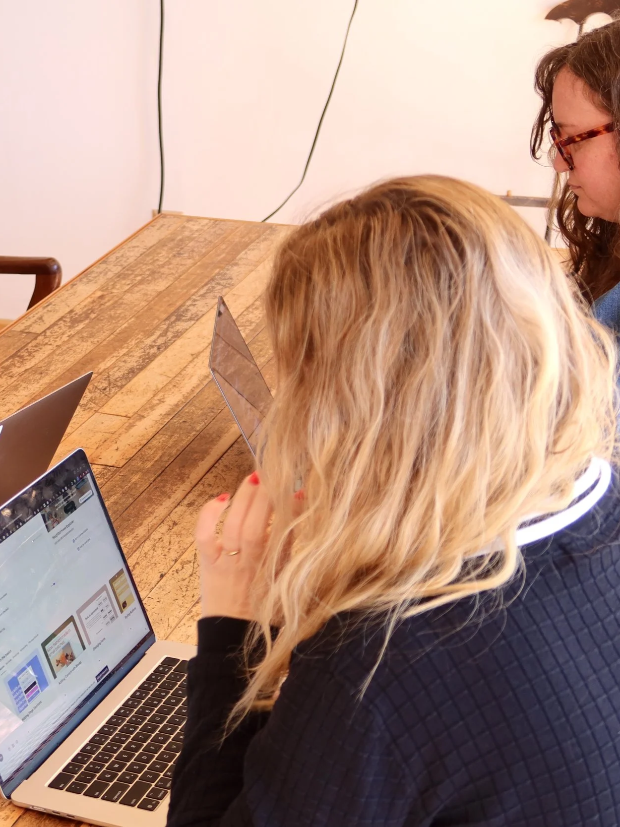 Two women sitting at a wooden table working on laptops and tablets.