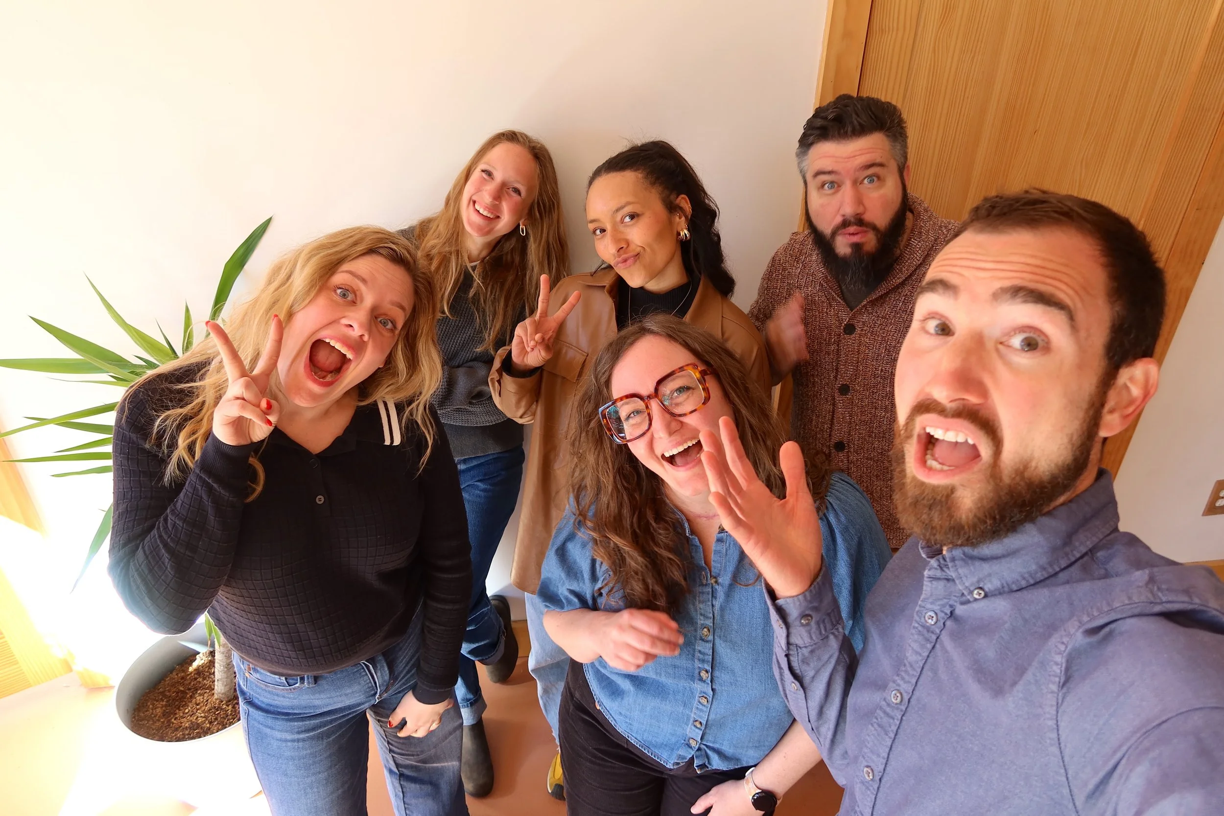 Group of six diverse adults taking a selfie together indoors, smiling and making silly faces and gestures.