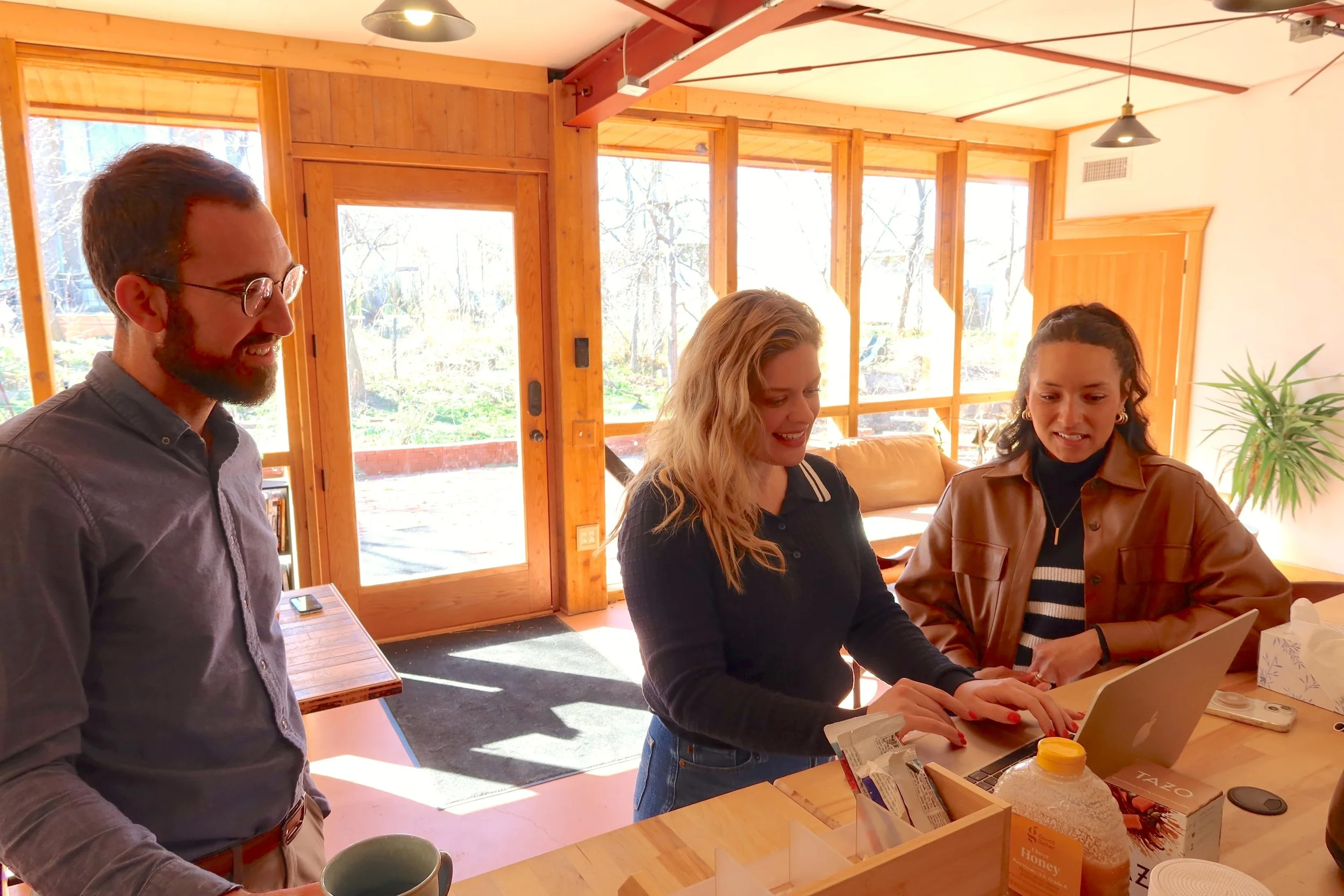 Three people gathered around a counter, looking at a laptop and smiling inside a bright, wooden-finished room with large windows.