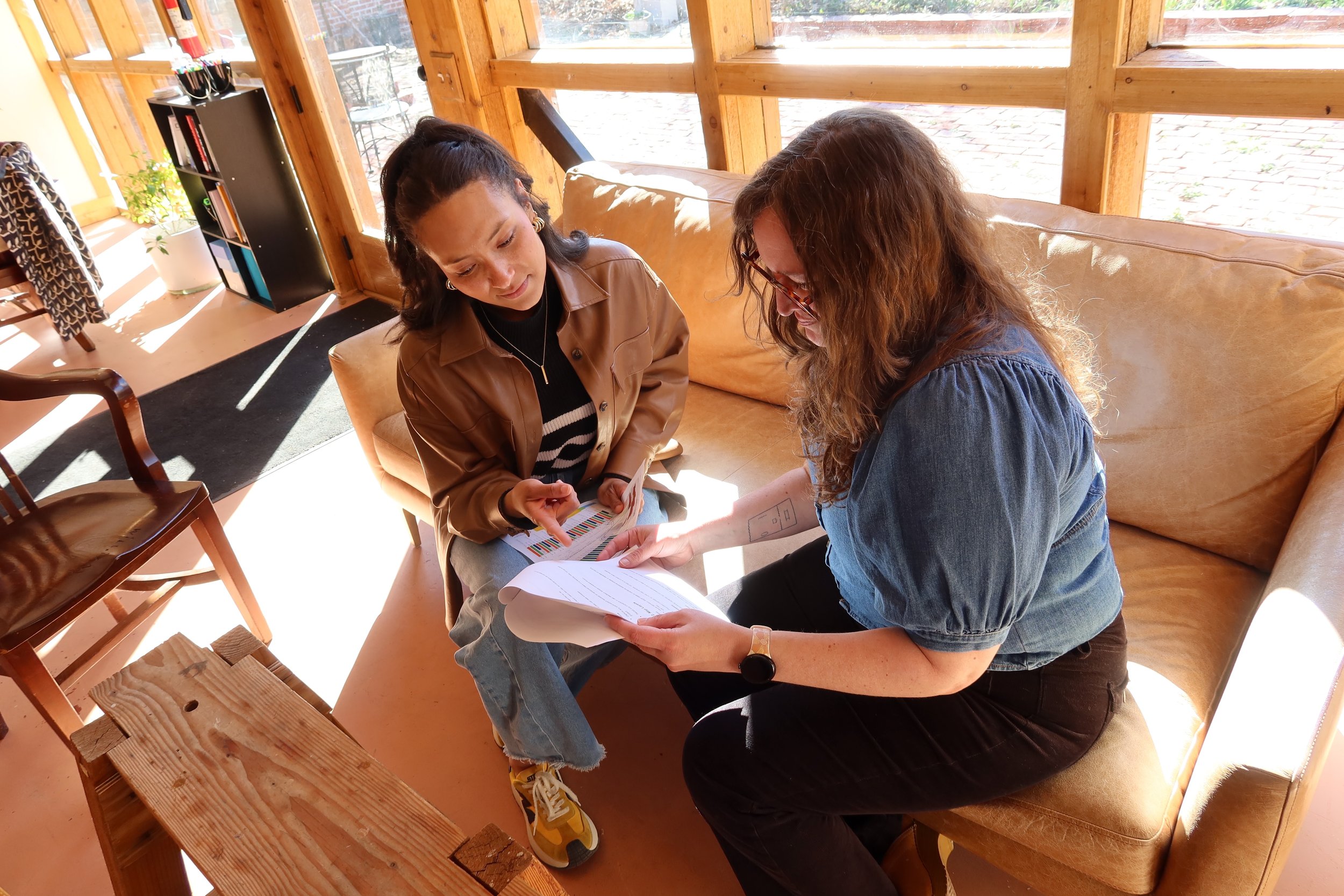 Two women sitting on a beige sofa, engaged in a discussion while looking at papers and a colorful chart, near large windows letting in sunlight.