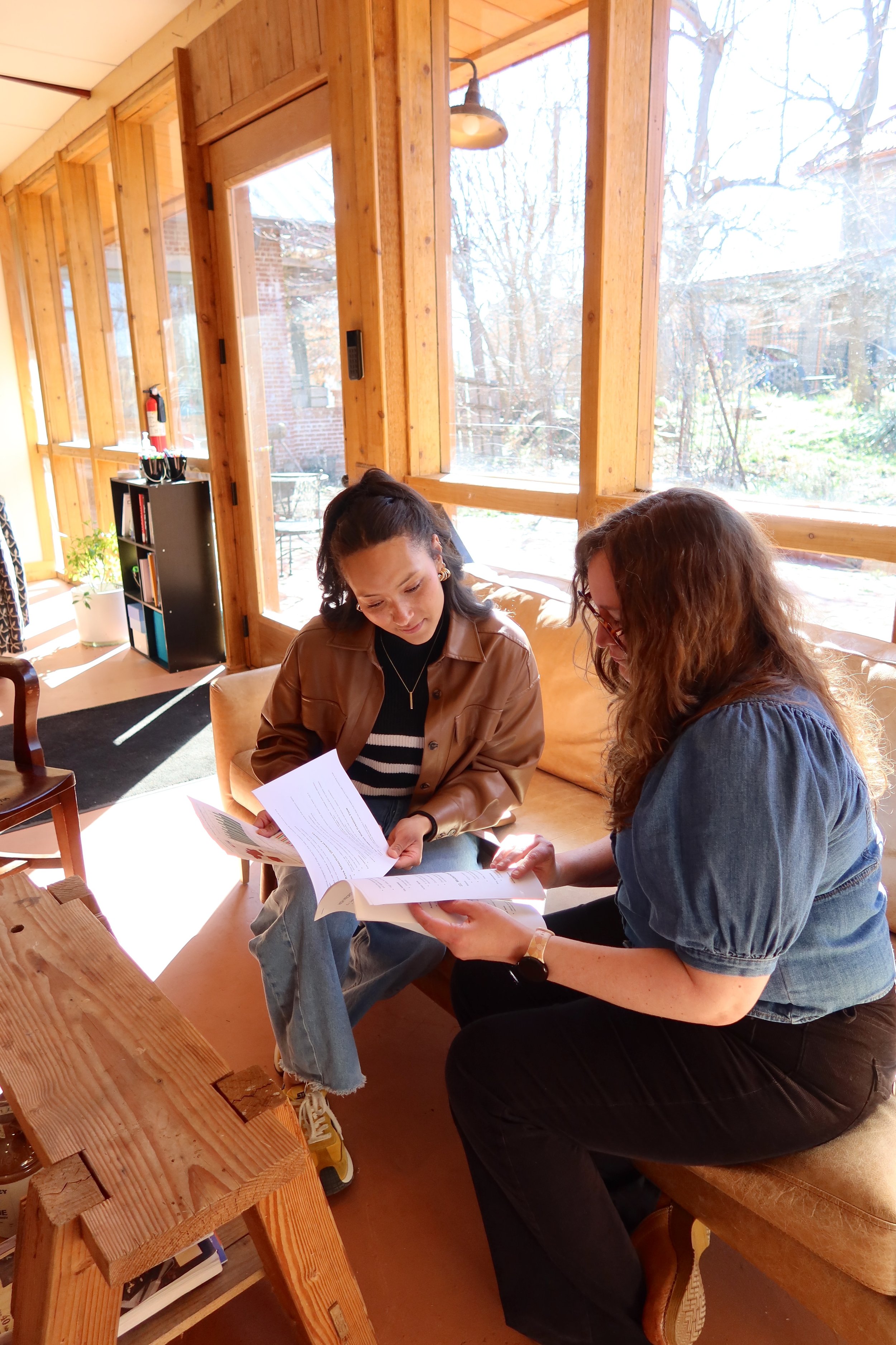 Two women sitting on a beige couch by large windows, reading documents together in a sunlit room with wooden walls.