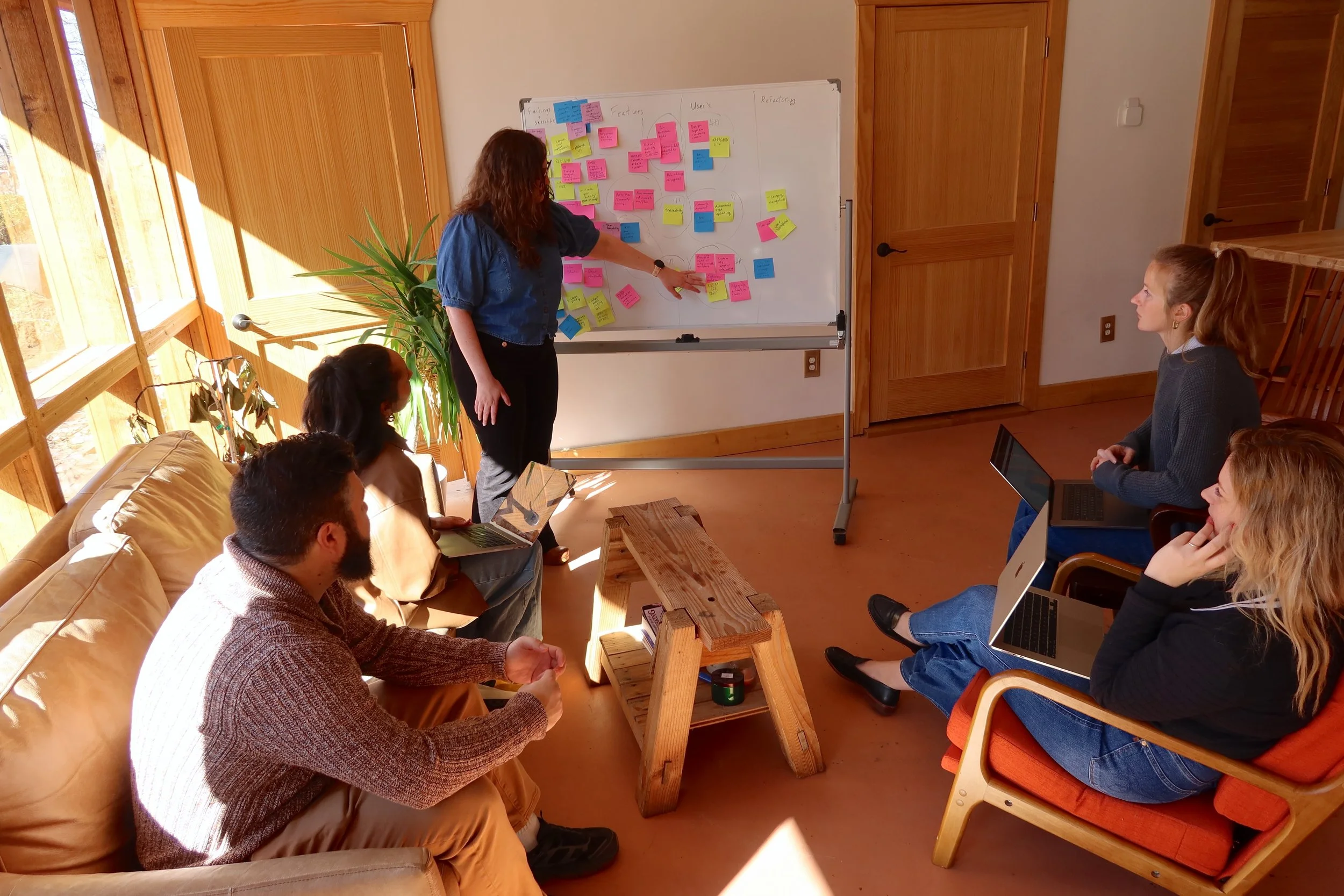 A woman is standing and pointing at a whiteboard covered with colorful sticky notes, while five people sit nearby in a cozy, sunlit room, some with laptops.