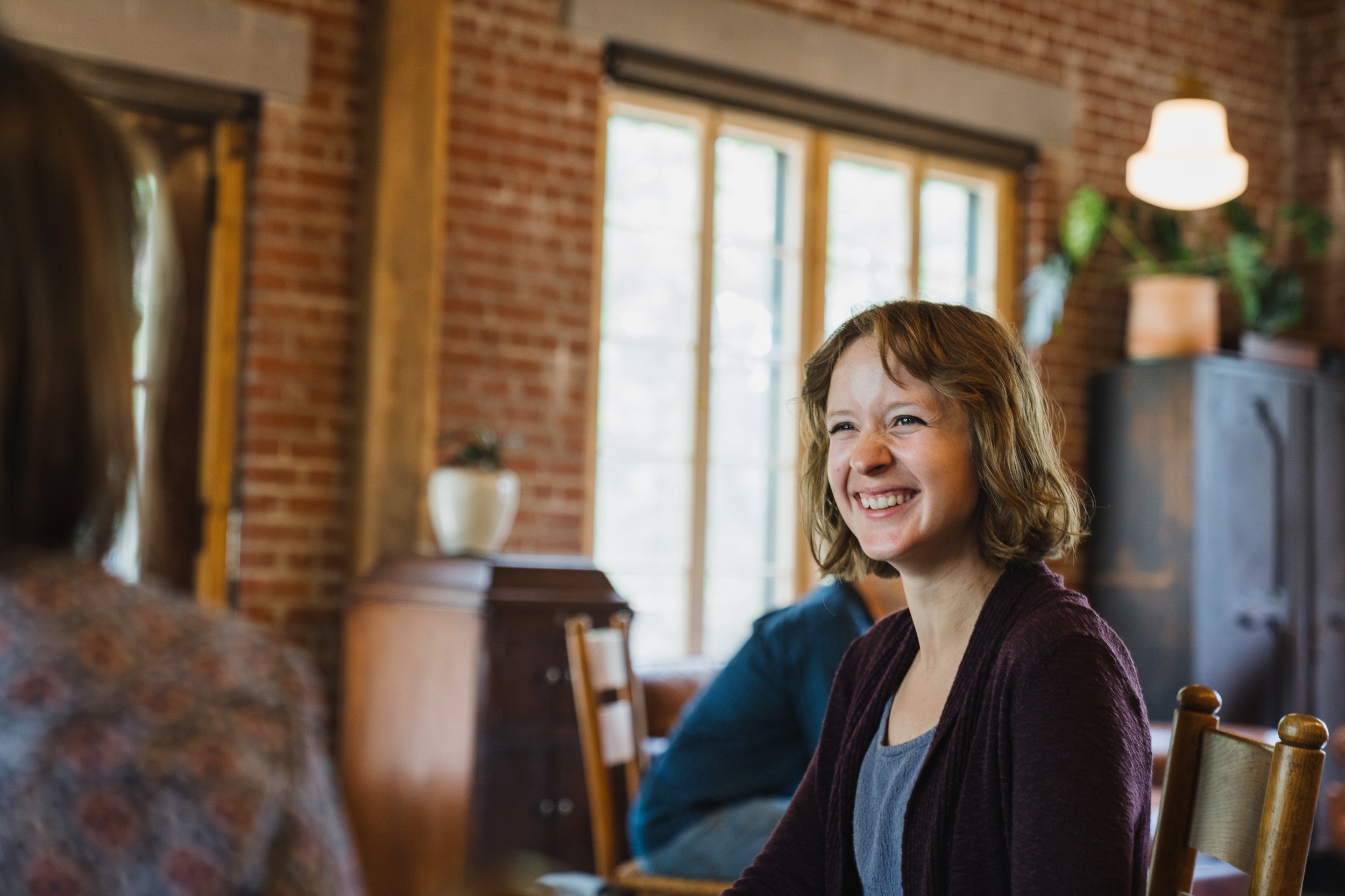 A woman with short brown hair smiling brightly in a cozy, rustic room with wooden and brick interior.