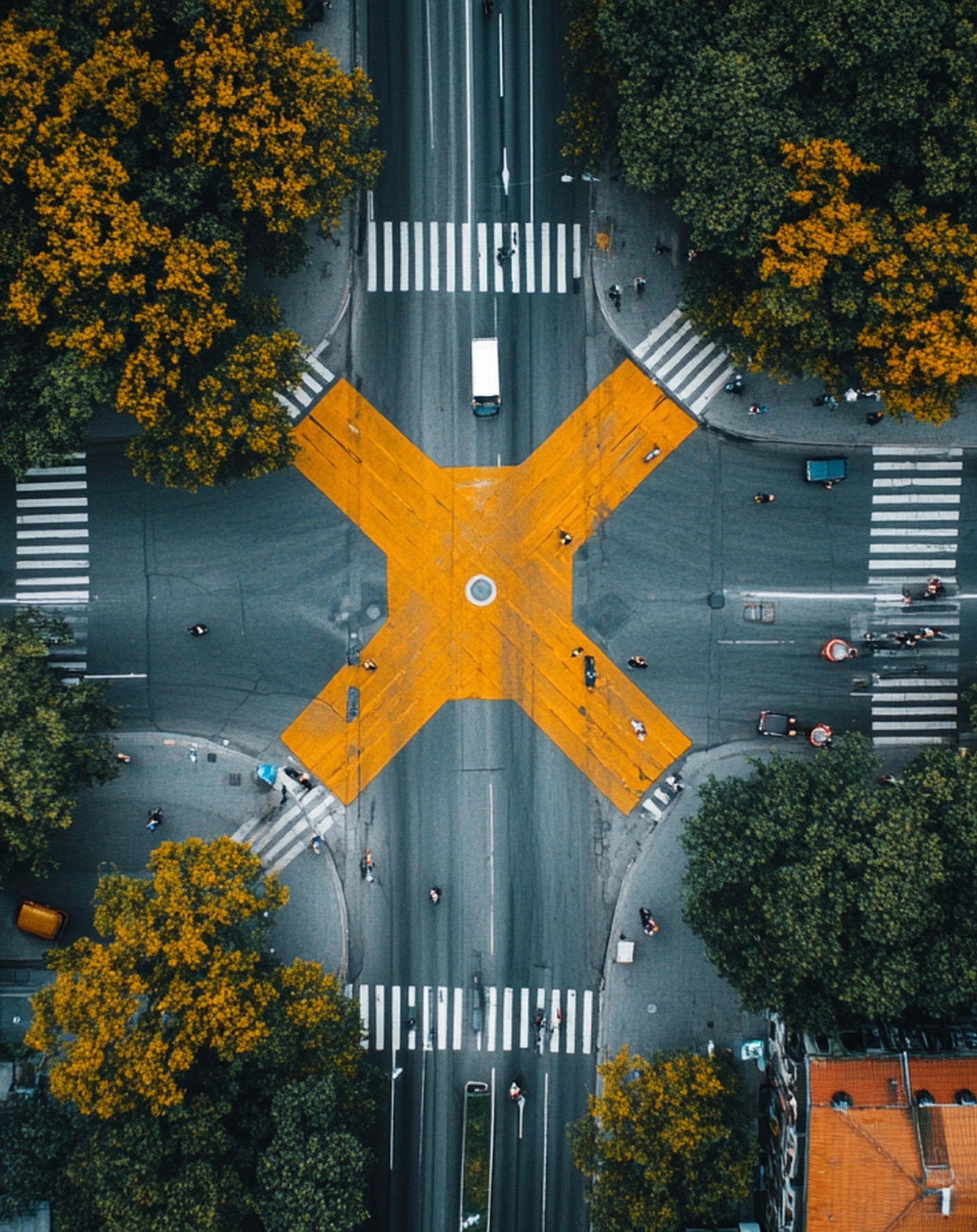 An aerial view of an intersection with four crosswalks, a yellow X road marking, and surrounded by trees with green and yellow leaves.