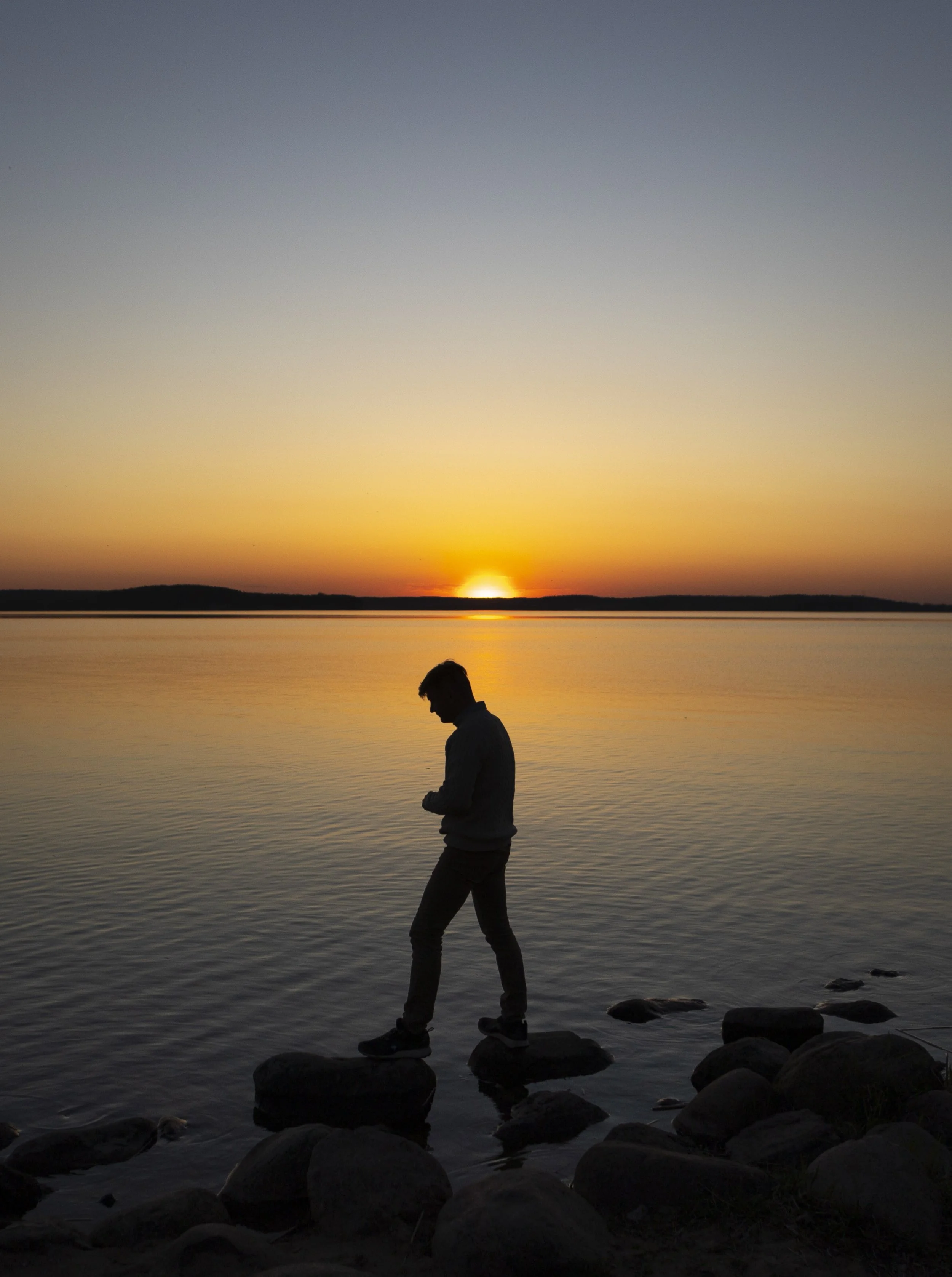 A person standing on rocks by the water at sunset, looking down, with a sunset over a calm lake in the background.