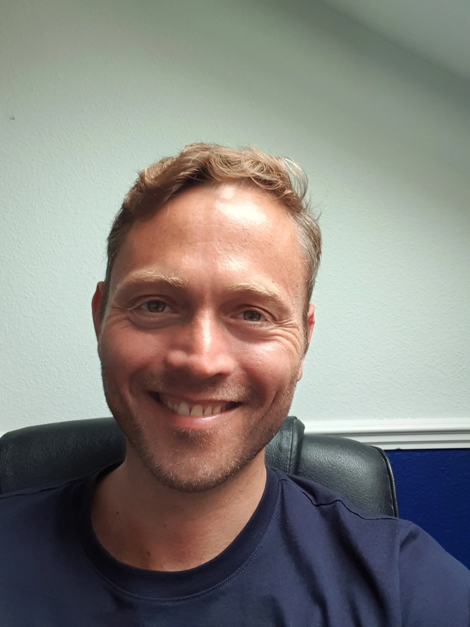 A smiling man with short light brown hair and a beard, sitting in a black office chair against a light-colored wall.