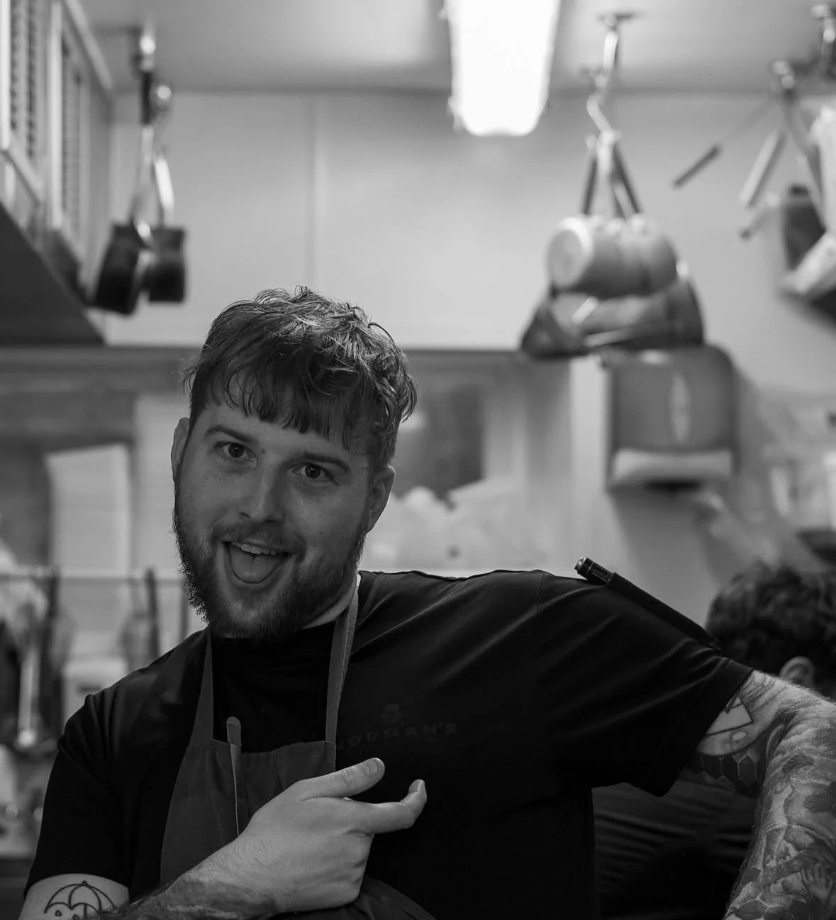 A young man with a beard and tattoos pointing to his shoulder in a kitchen or workshop setting, with various tools and equipment hanging on the walls.
