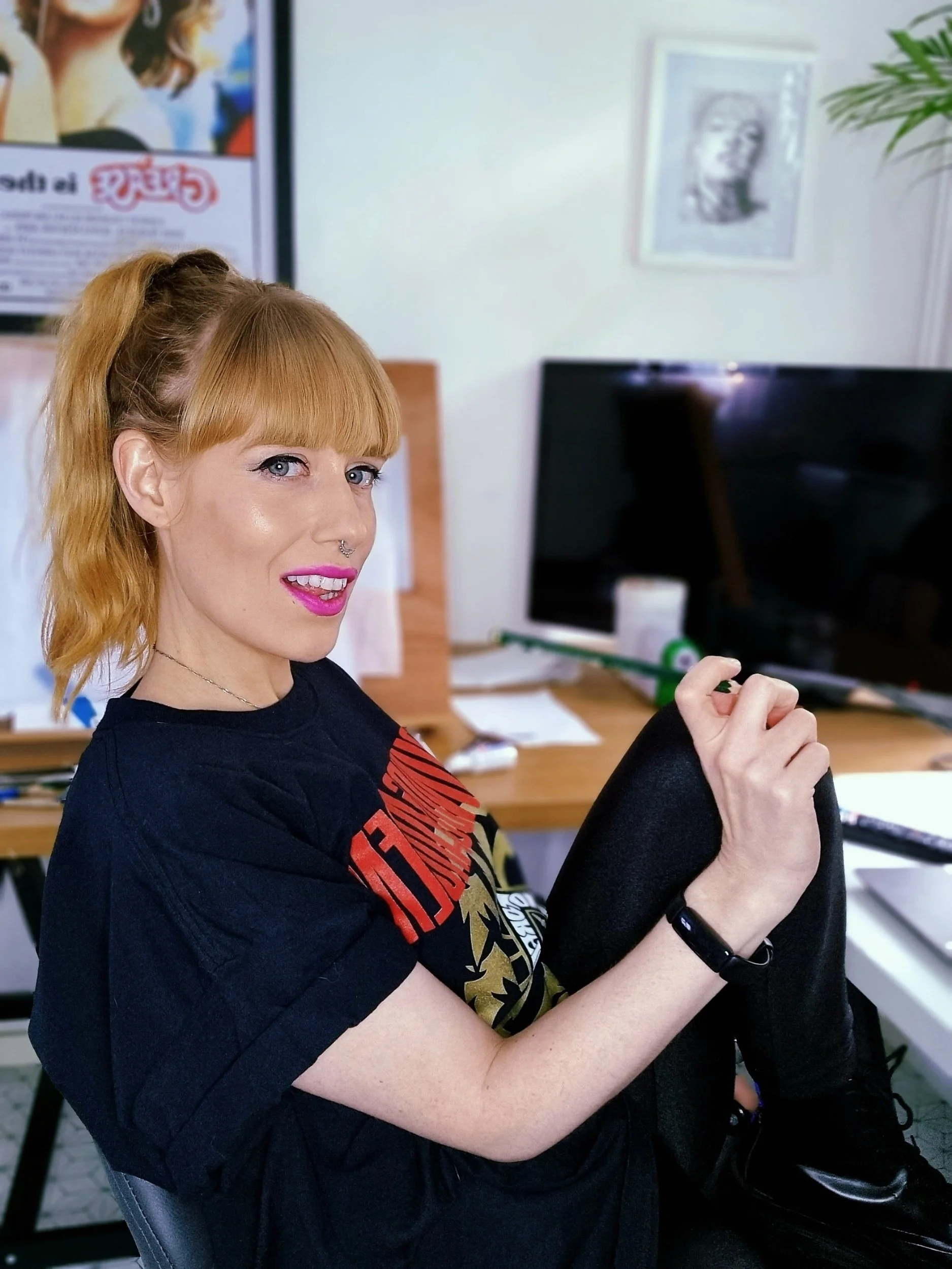 A woman with red hair, wearing a black graphic T-shirt and black pants, sitting indoors with a computer monitor and various objects on a desk behind her.