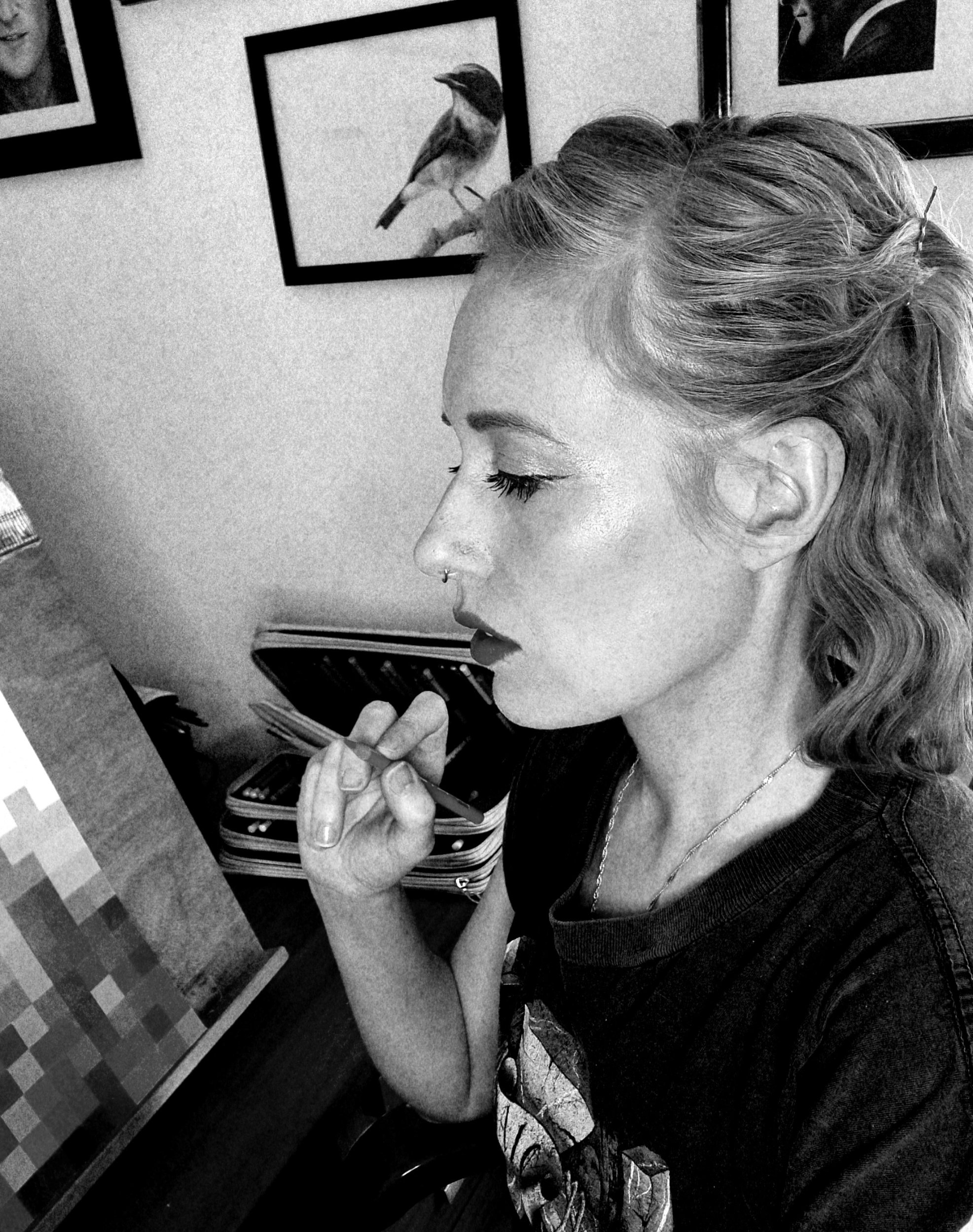 A woman with wavy hair and a nose piercing sitting at a desk, looking at her laptop. Behind her, framed bird photographs are on the wall.