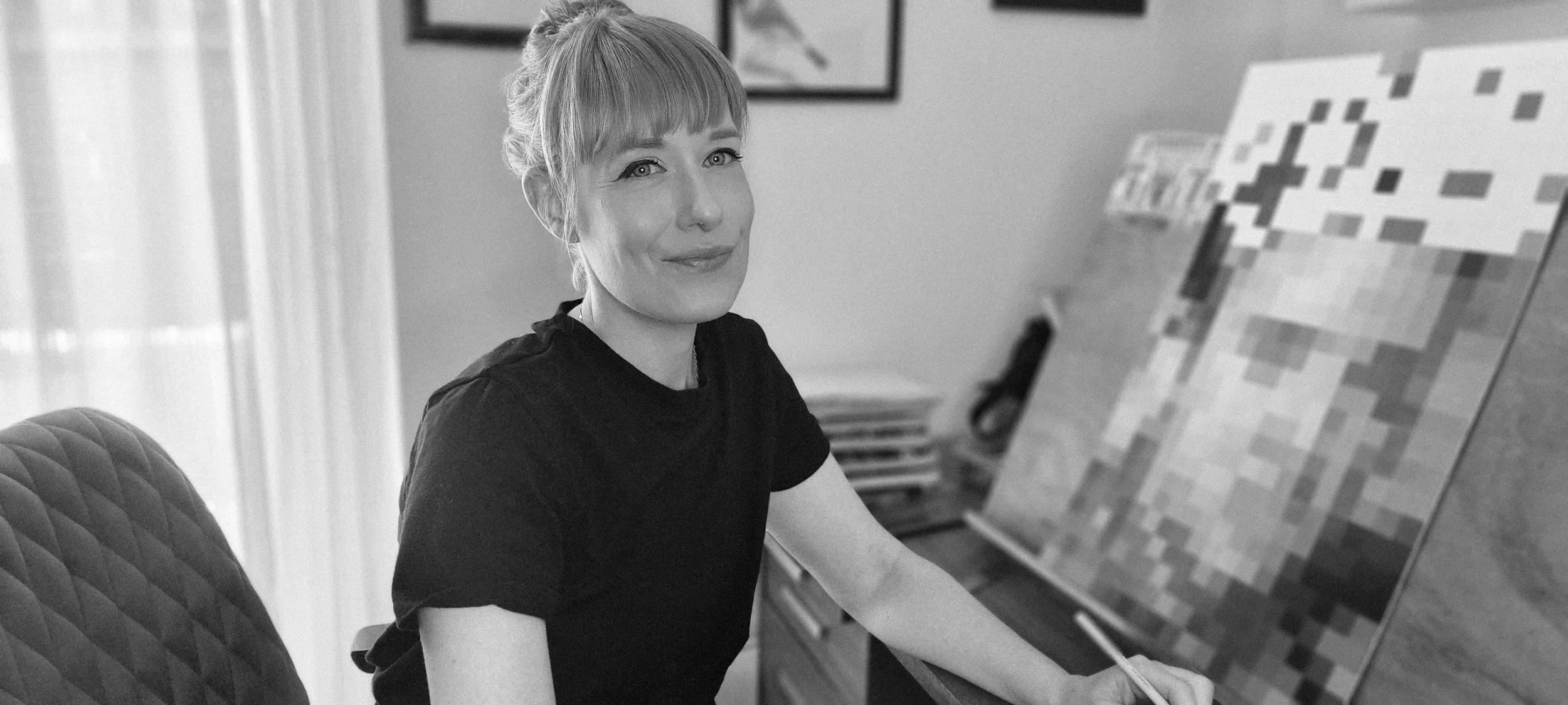 A woman with light hair and a nose piercing, wearing a black t-shirt, sits at a desk with a pixel art painting of a character from Minecraft on an easel. She is looking at the camera and smiling.