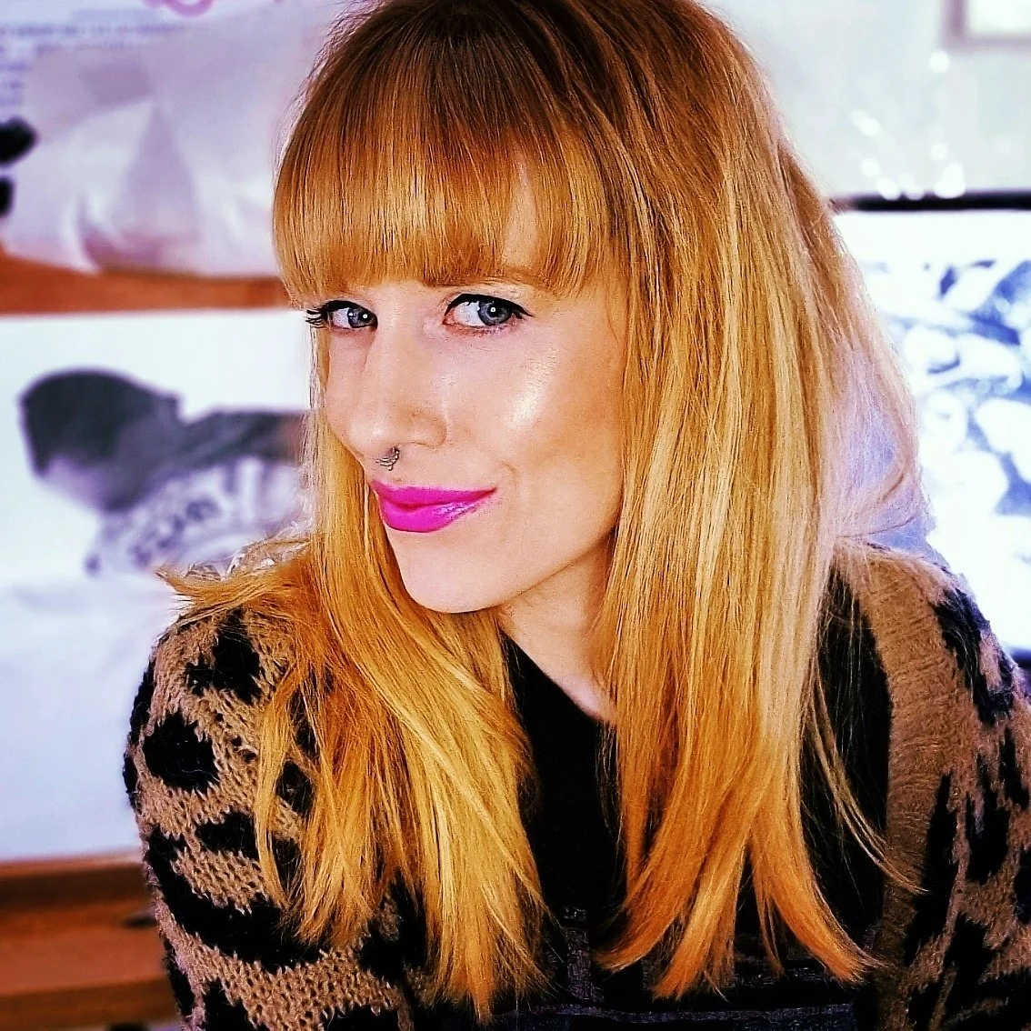 A woman with long, red hair and bangs, wearing bright pink lipstick, a nose septum piercing, and a black and beige animal print sweater, smiling at the camera.