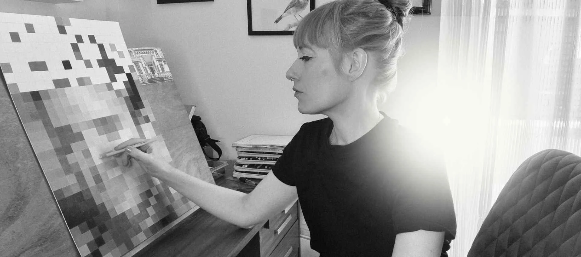 A woman in a black shirt sits at a desk, using a stylus to work on a pixelated mosaic art piece on a canvas, with sunlight coming through a window behind her.
