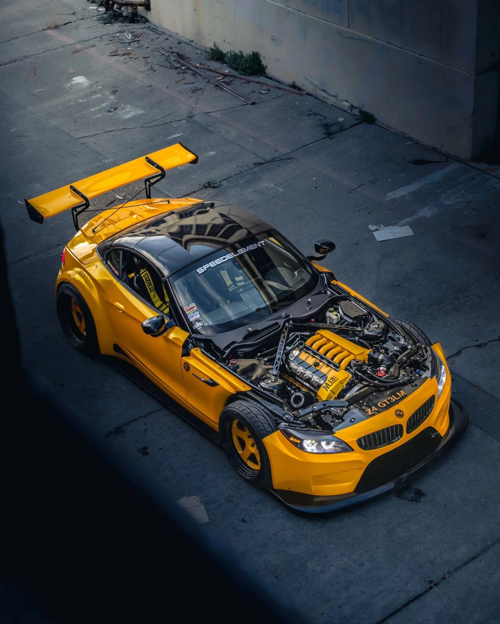 A yellow sports car with a black hood and roof, a large rear wing, and racing decals, parked on a concrete surface with an open hood showing the engine.