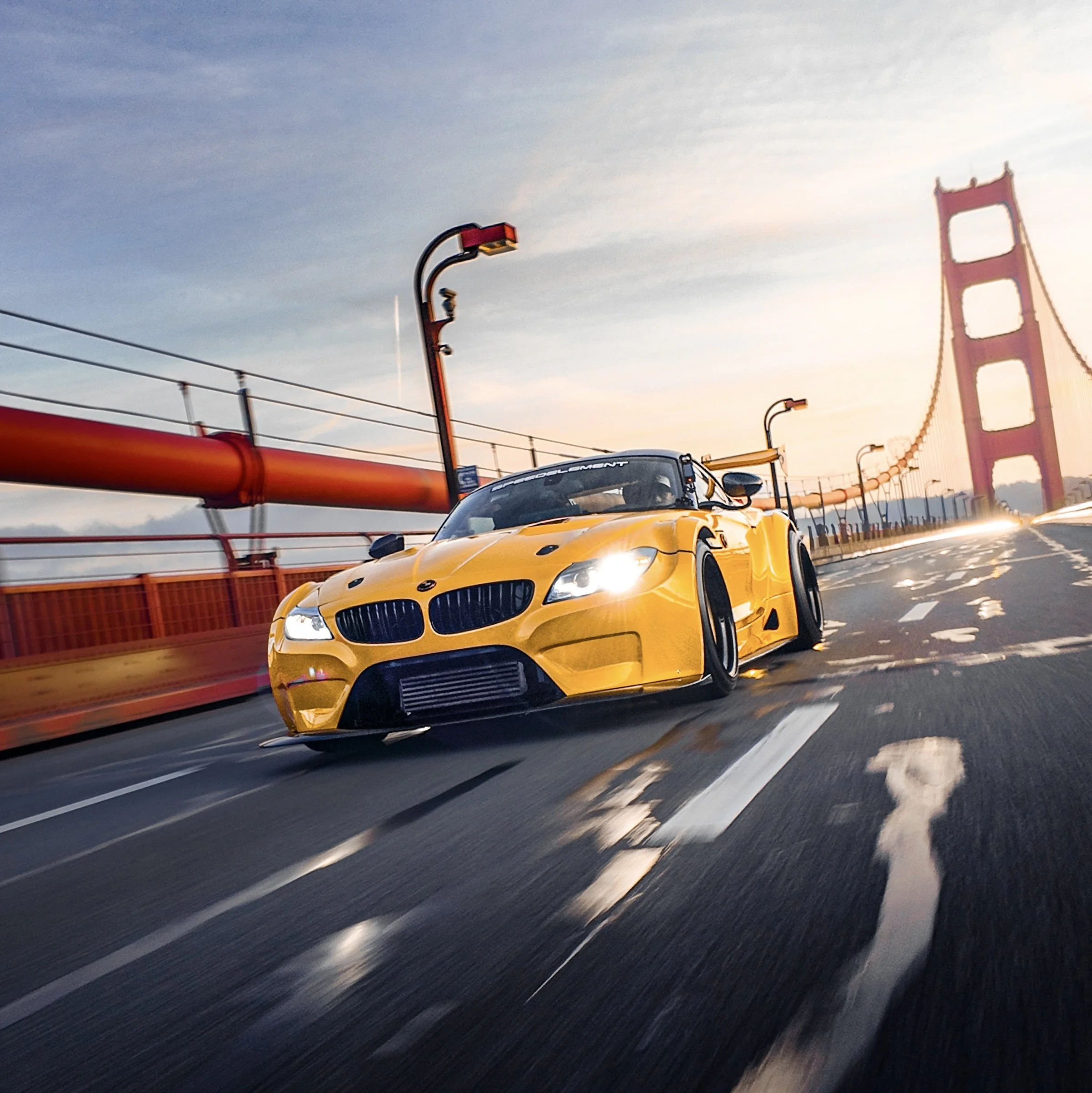 A yellow racing car driving on a bridge with the San Francisco Golden Gate Bridge in the background during sunset.