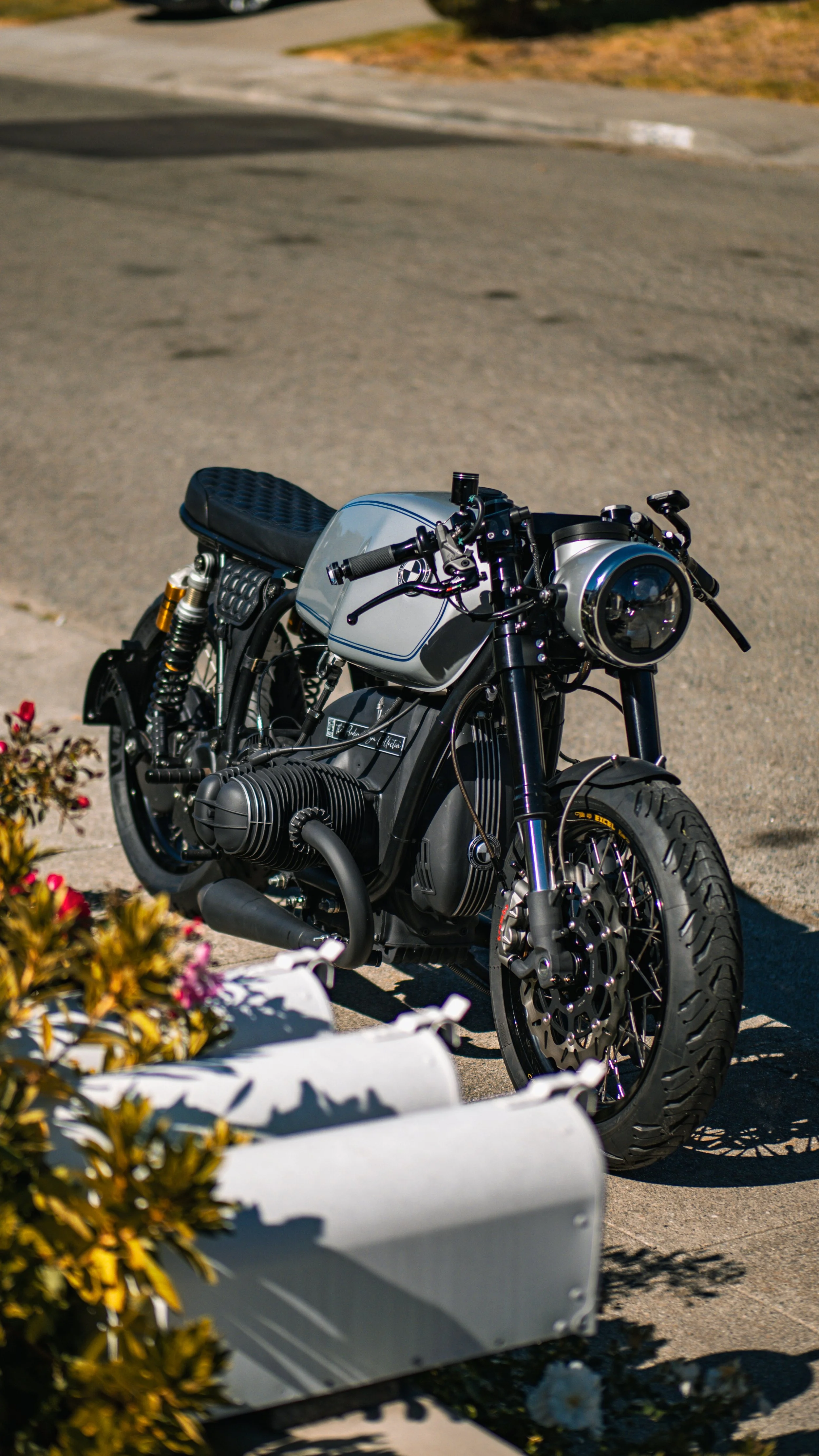 A modern custom black motorcycle parked on a sidewalk with plants in the foreground, a road and grass in the background during daytime.