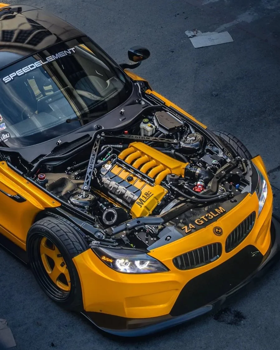 Yellow sports car with its hood open, revealing a detailed engine. The windshield has a decal that says 'SPEEDELEMENT.' The car has yellow wheels and tires, and the front bumper is black. The engine has yellow components and is labeled with 'M18' and 'Z4 GT3 LM.'