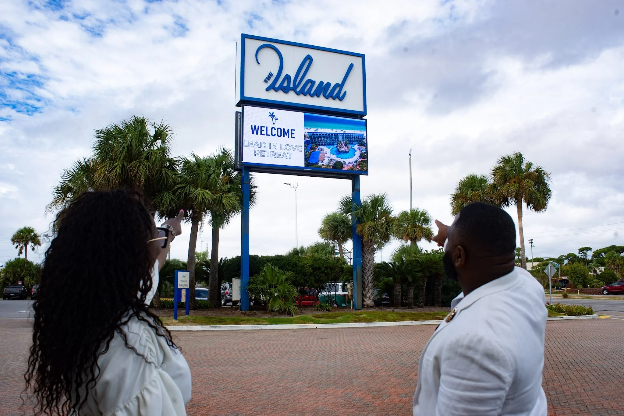 Two people standing outdoors in front of a large sign that reads 'The Island' and a digital display that says 'Welcome Lead in Love Retreat', with palm trees and a cloudy sky in the background.