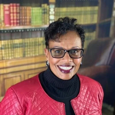 A woman with short curly hair, glasses, and earrings smiling, wearing a red quilted jacket and black turtleneck in front of a bookshelf filled with colorful books.