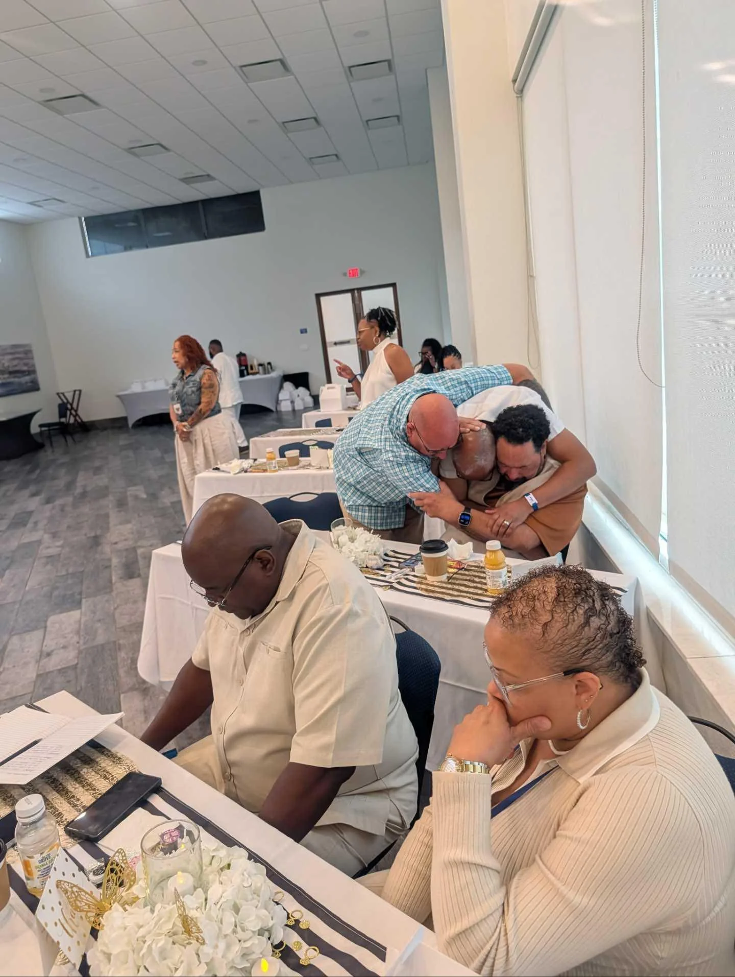 People gathered in a social setting, some seated at tables with drinks and decorations, others standing and engaging in conversations, in a well-lit room with large windows and a high ceiling.