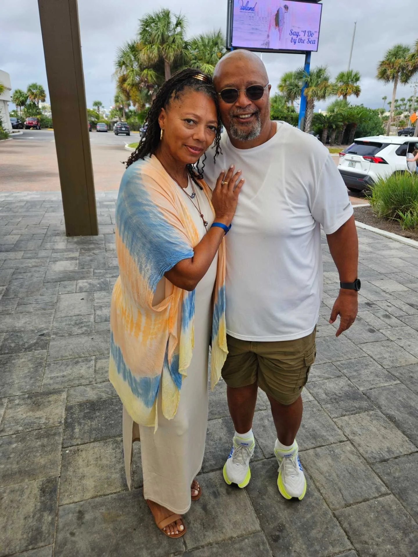 A woman and a man are standing close together outdoors, smiling at the camera. The woman is wearing a pastel-colored, tie-dye shawl and a cream dress, and the man is dressed in a white T-shirt, olive shorts, and white sneakers, with sunglasses and a watch. In the background, there are palm trees, parked cars, and a digital billboard.