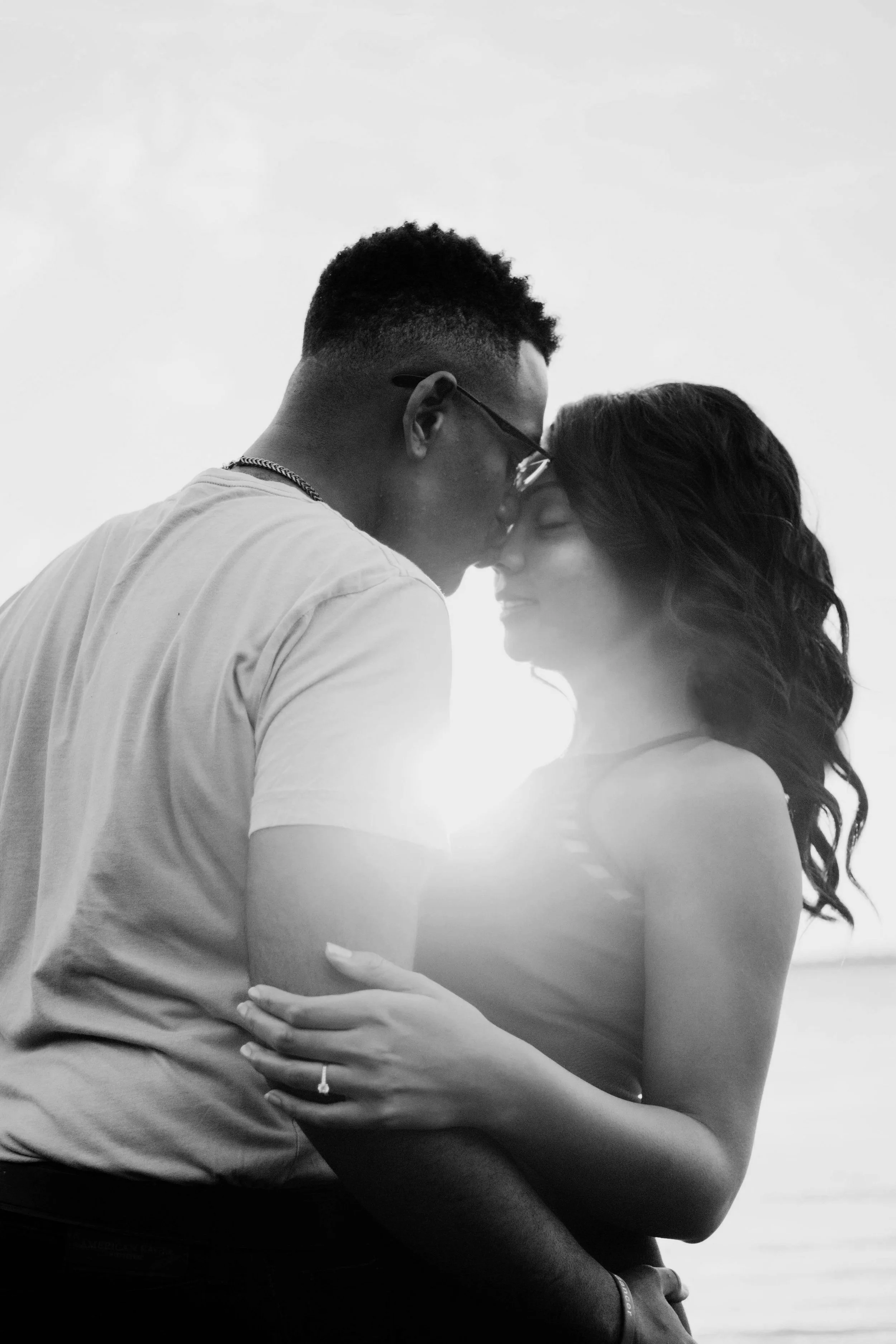 A black-and-white photo of a couple embracing closely, with foreheads touching and eyes closed, illuminated by sunlight behind them.
